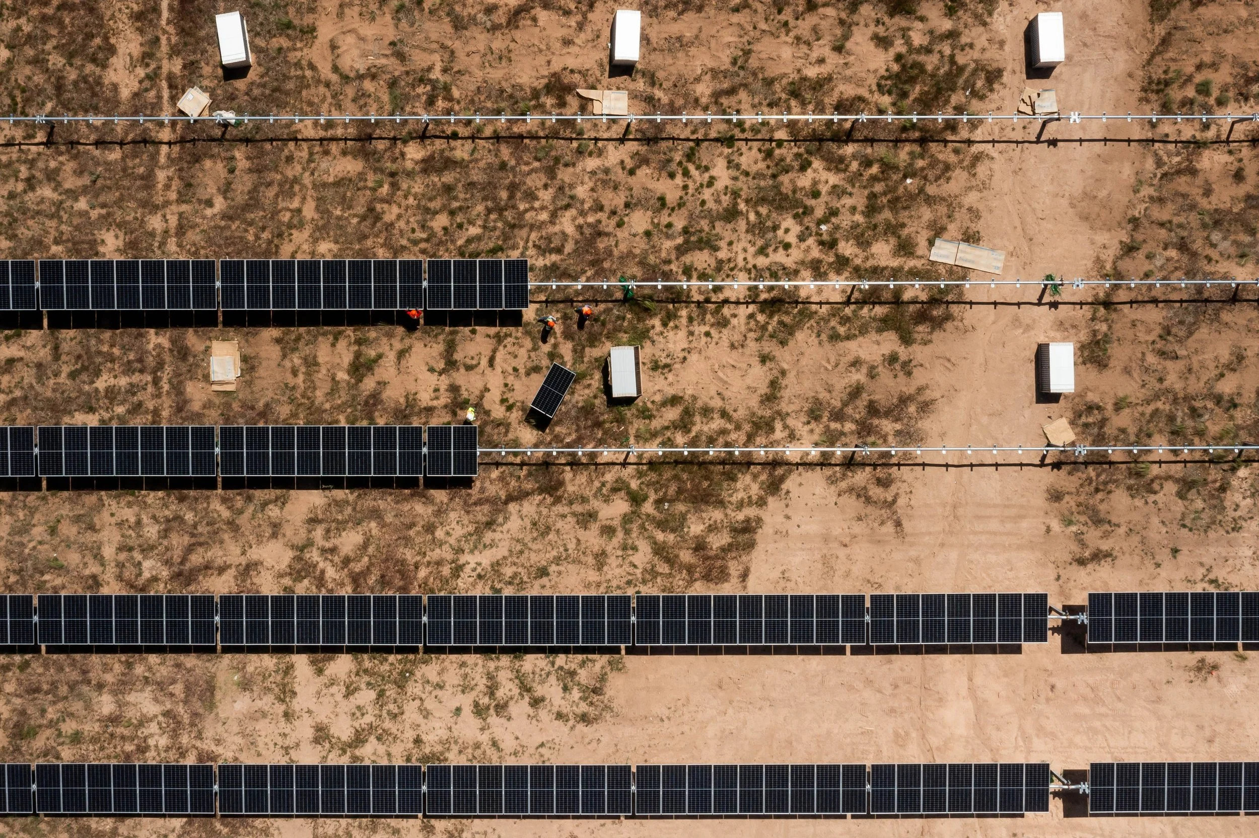 Aerial view of solar panels arranged in rows in a desert, with workers installing some panels and scattered small structures surrounding the panels.