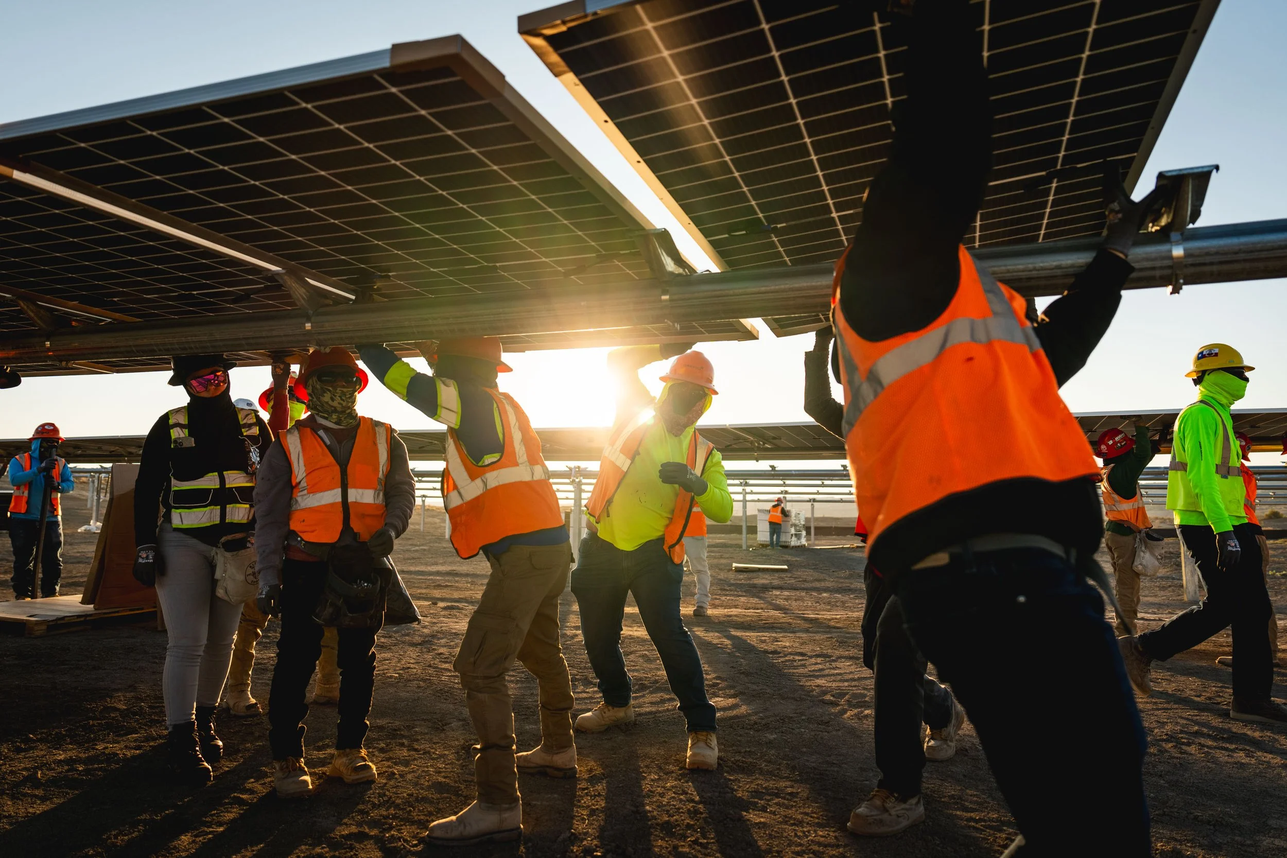 Workers installing solar panels outdoors at sunset, wearing safety vests, helmets, and masks.