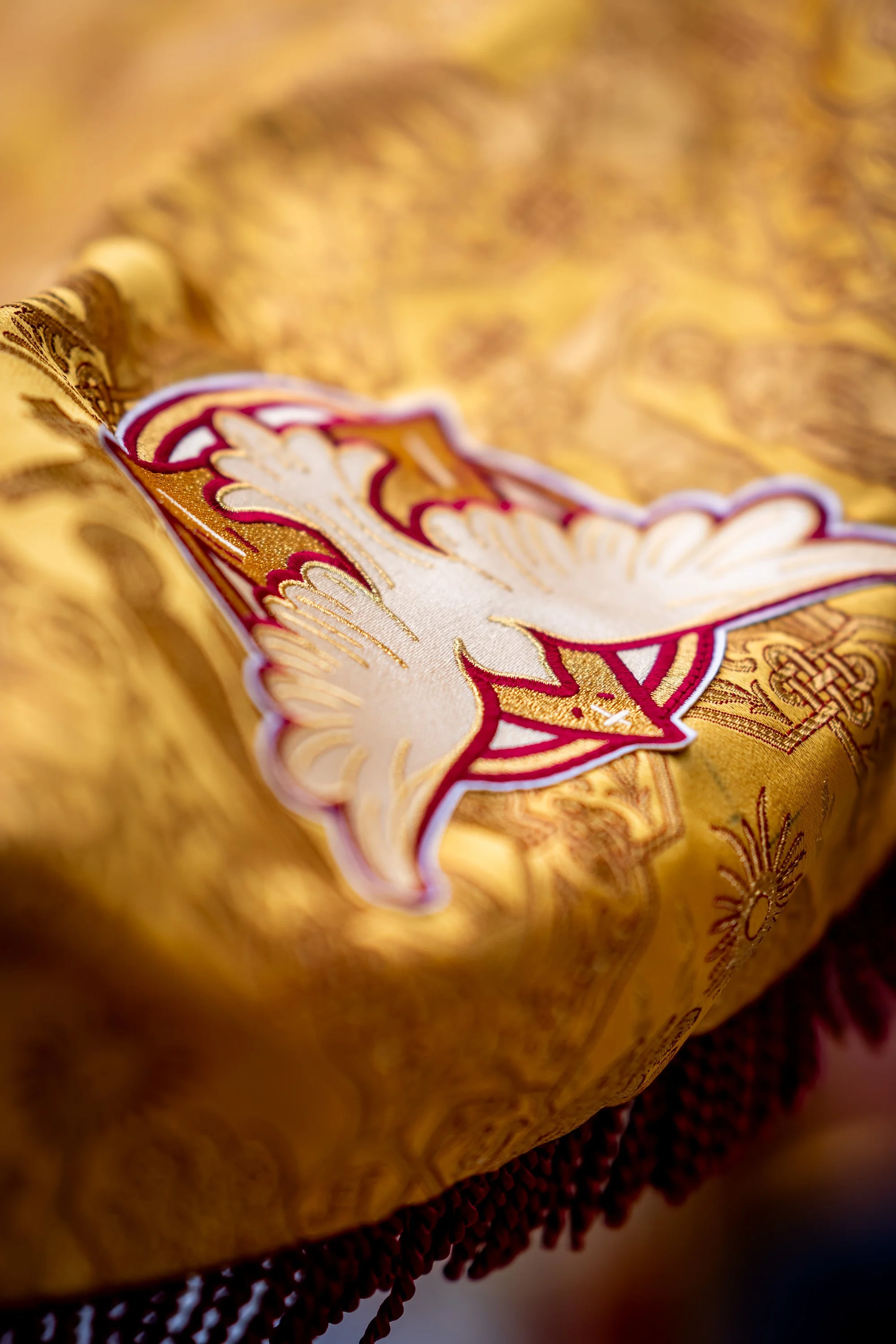 Close-up of a decorative cloth with an embroidered dove design, featuring gold, white, red, and purple colors, on a golden patterned fabric background.