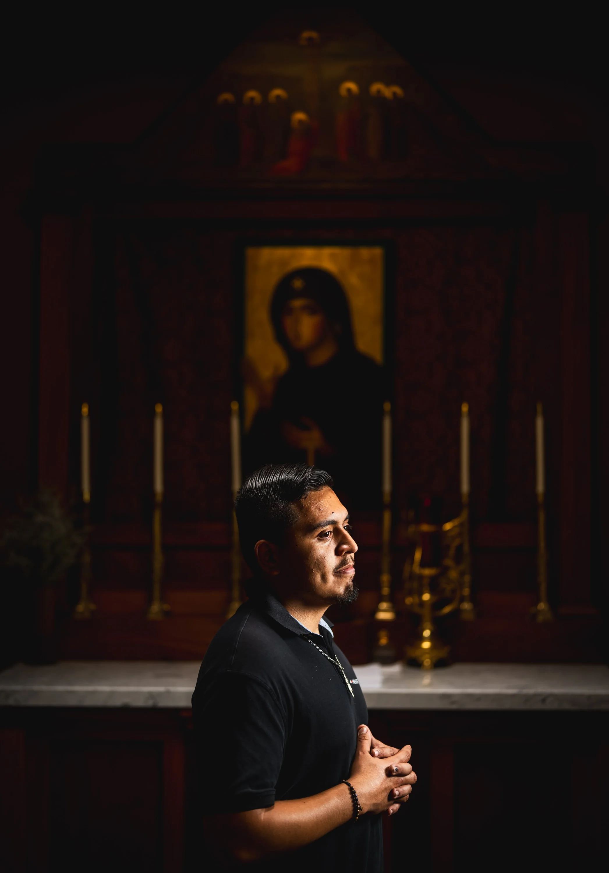 A young man standing with hands clasped in prayer inside a church, lit by soft light with an icon of a saint in the background.