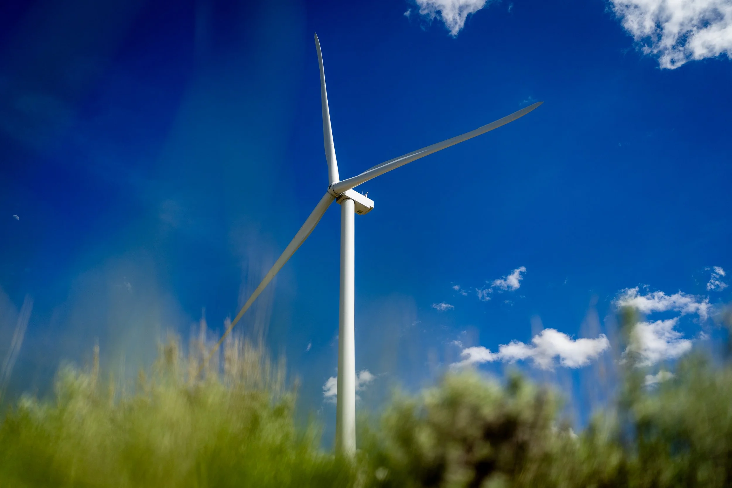 A large white wind turbine stands tall against a bright blue sky with some scattered clouds and a faint moon visible in the distance.