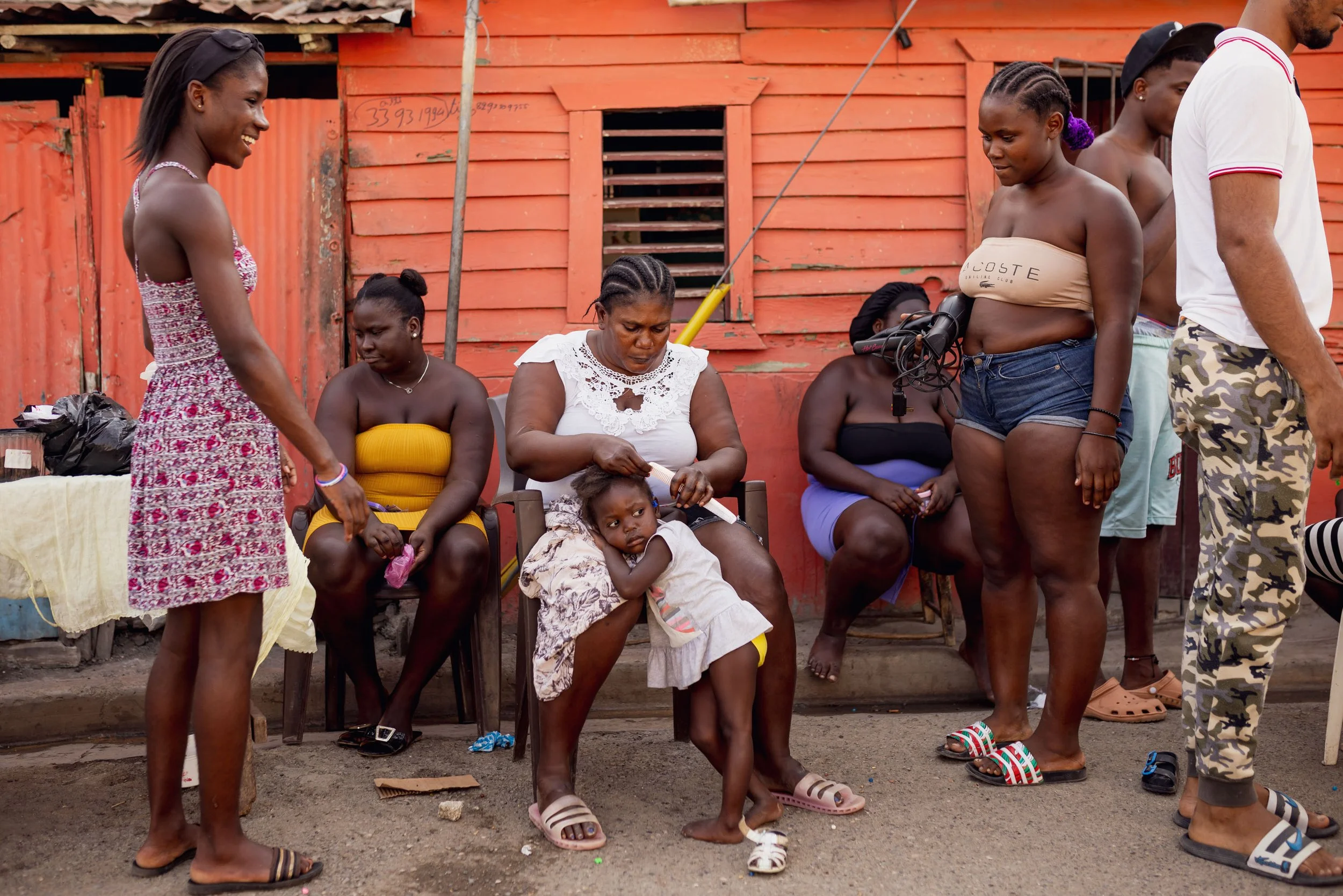 Group of women and children sitting and standing outside against a red wooden building, engaging in conversation.