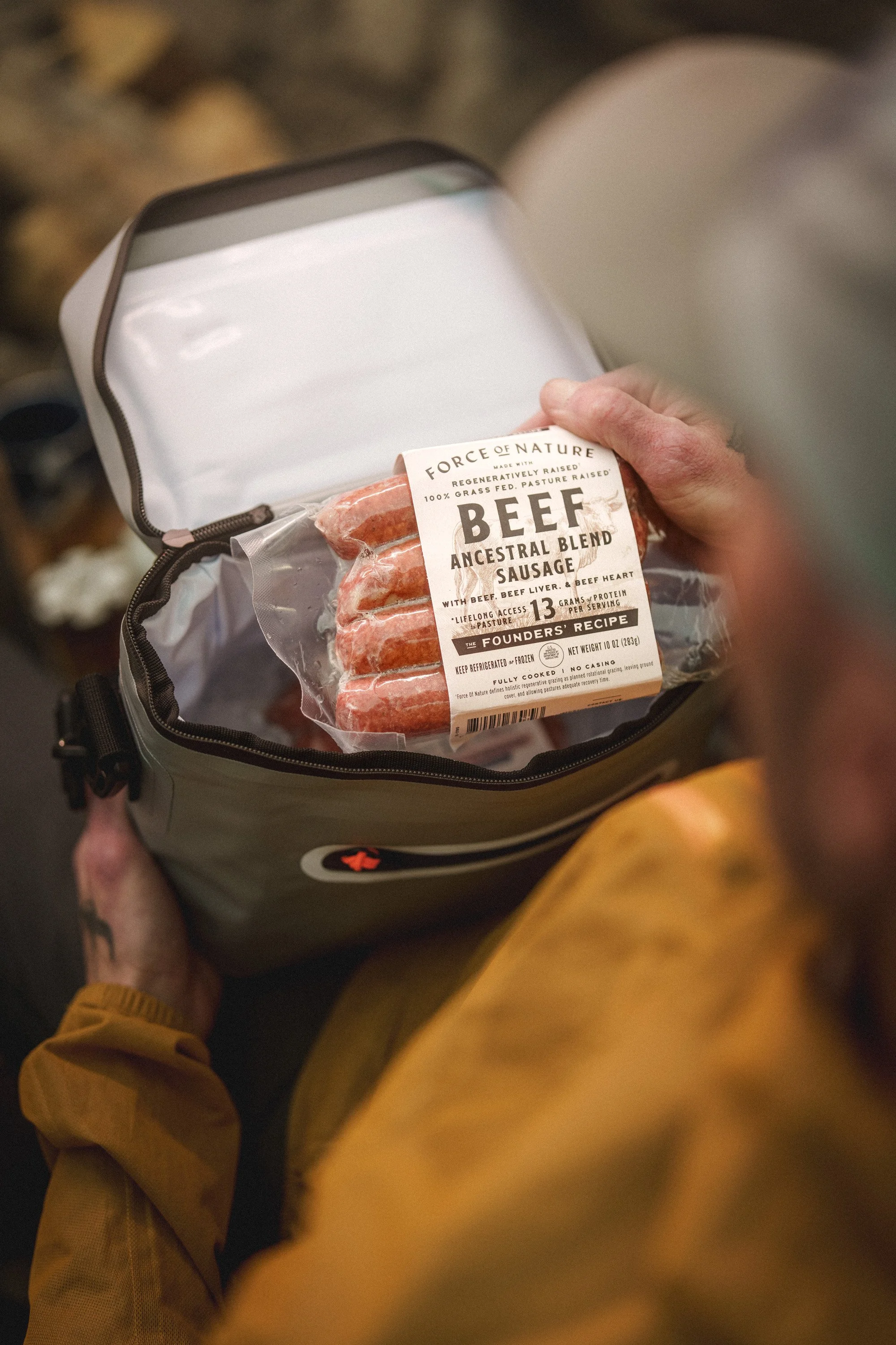 A person holding an open gray insulated bag with a pack of raw beef sausages inside, with the person's hand touching the packaging.
