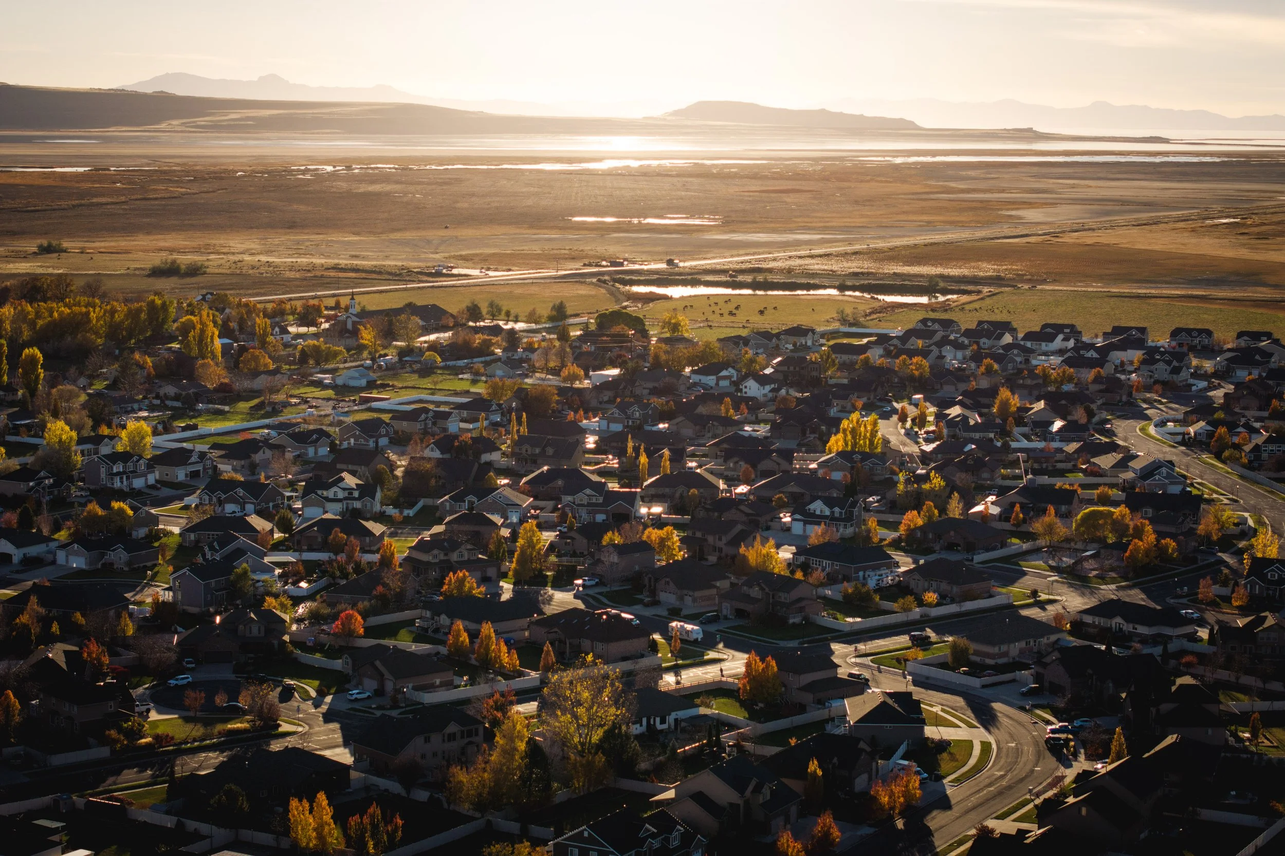 An aerial view of a suburban neighborhood with houses, trees, and winding streets at sunset, with mountains and open land in the background.
