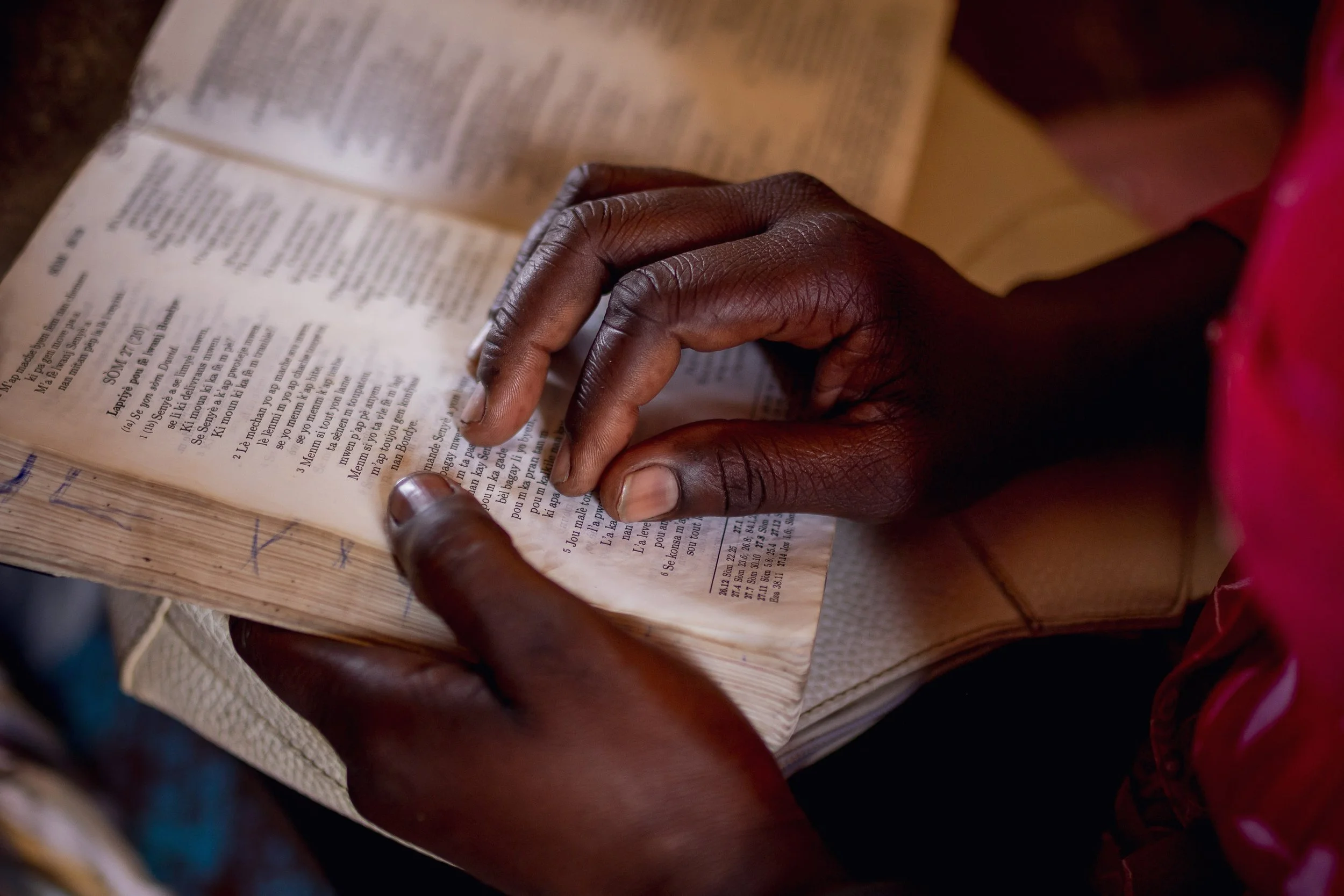 Close-up of a person's hands holding and reading an open book with text, with dark skin and wearing a red garment.
