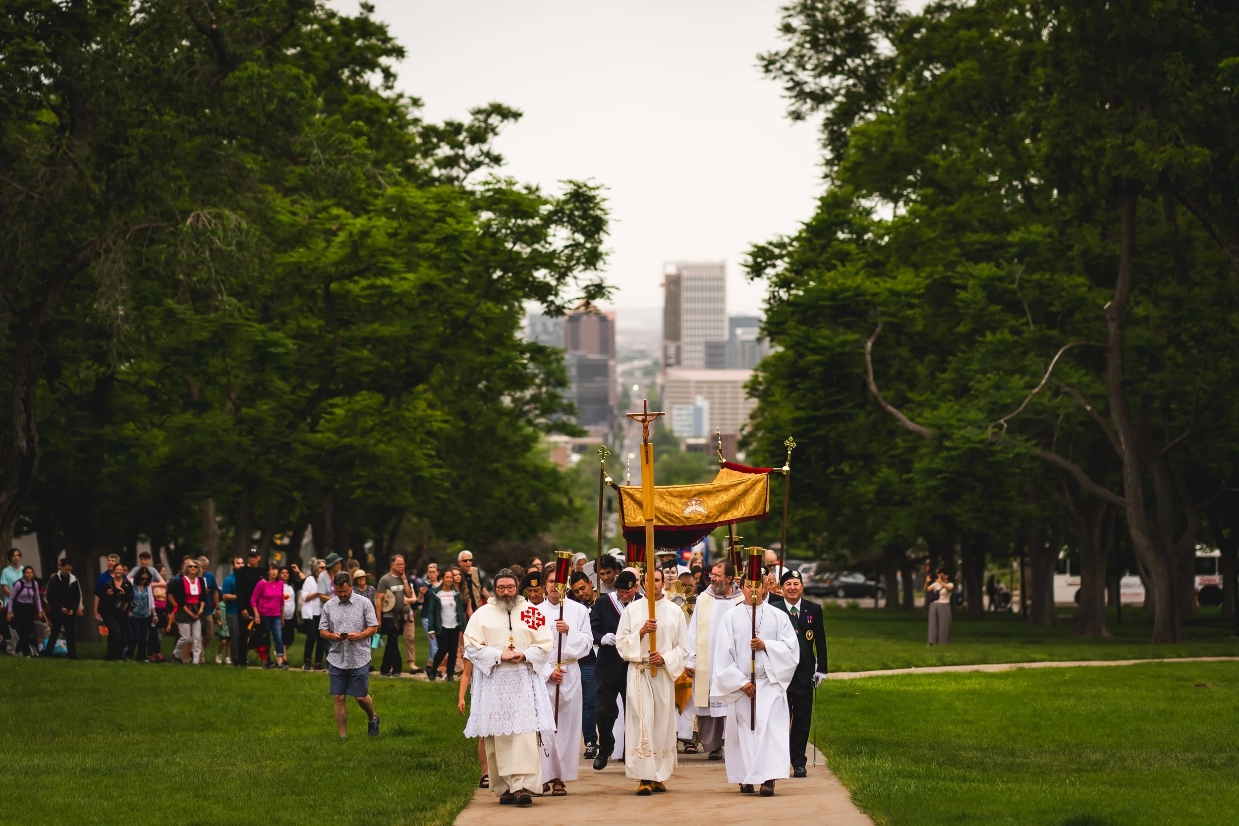 A religious procession of clergy and laity passes through a park with green trees and an urban skyline in the background, carrying a canopy and religious symbols.