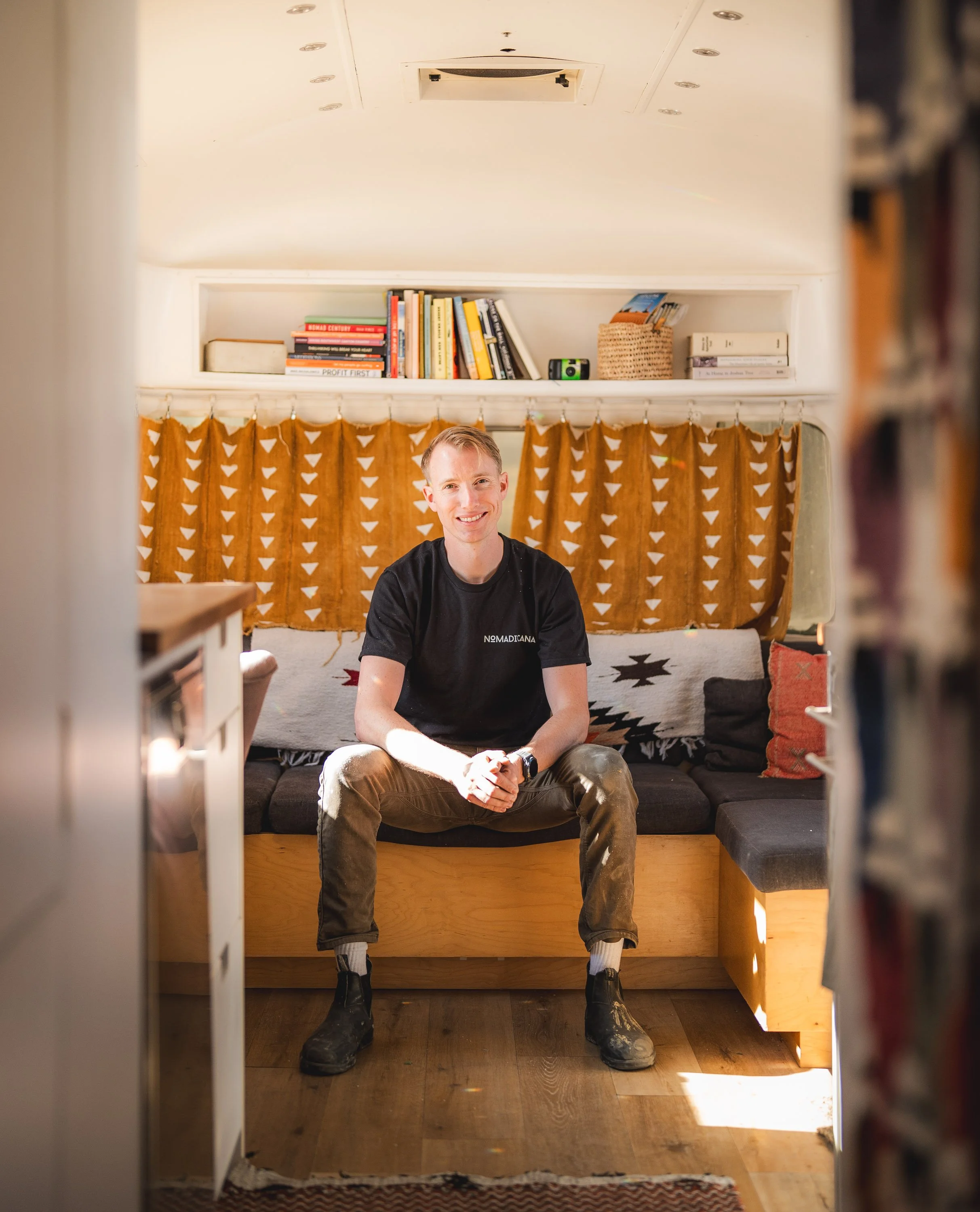 A man sits on a built-in window seat in a cozy room with orange curtains, bookshelves, and a colorful blanket behind him, smiling at the camera.