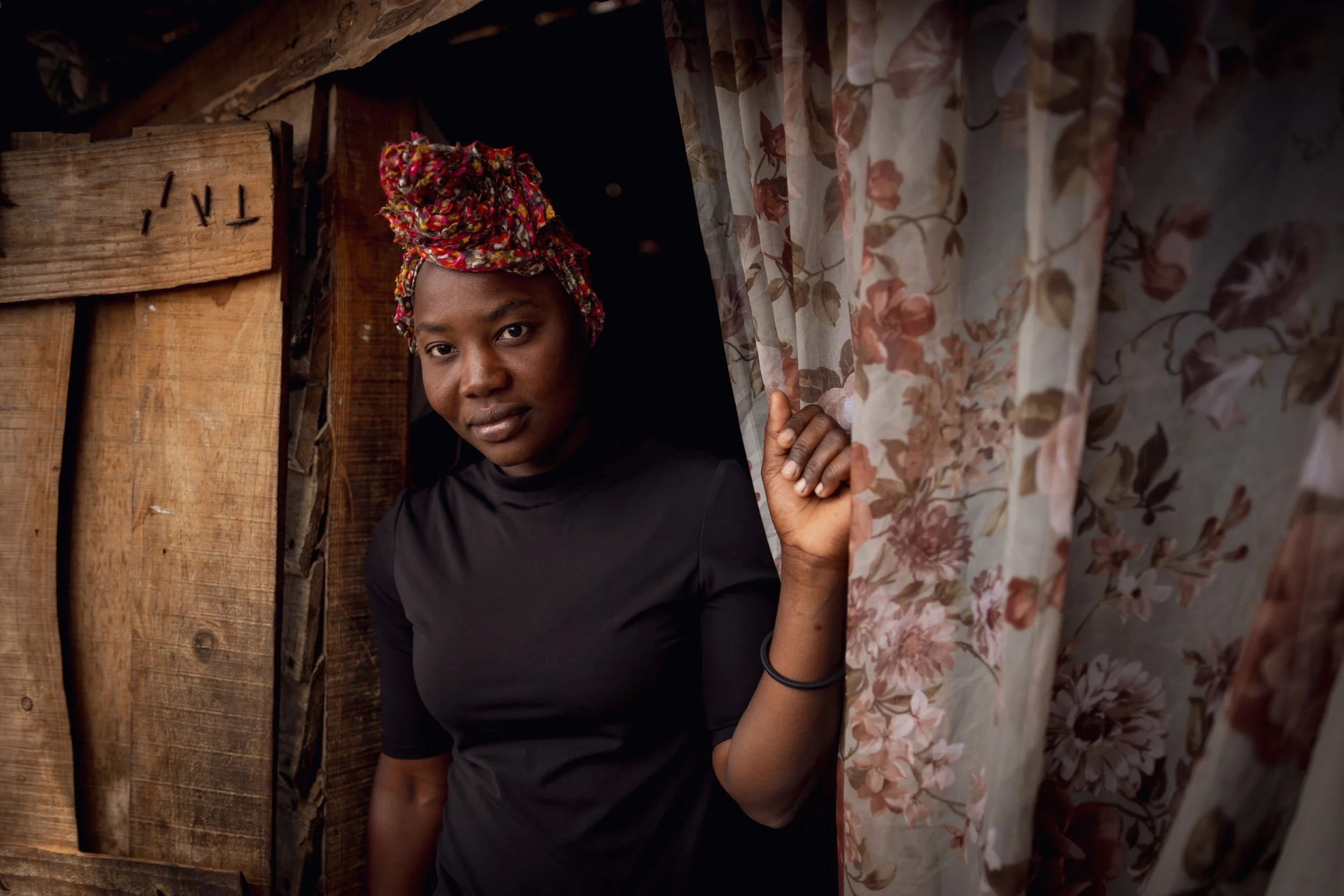 A woman with dark skin, wearing a black shirt and a colorful head wrap, peeks out from behind a floral curtain inside a wooden structure.