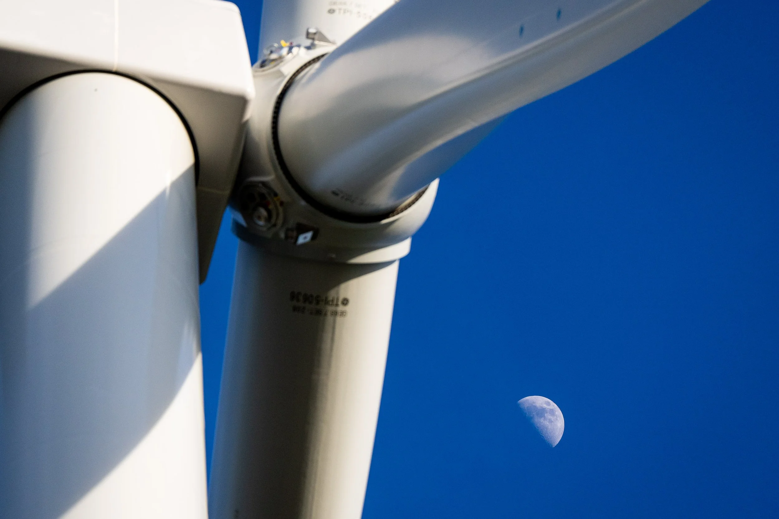 Close-up of a wind turbine with the moon visible in the clear blue sky