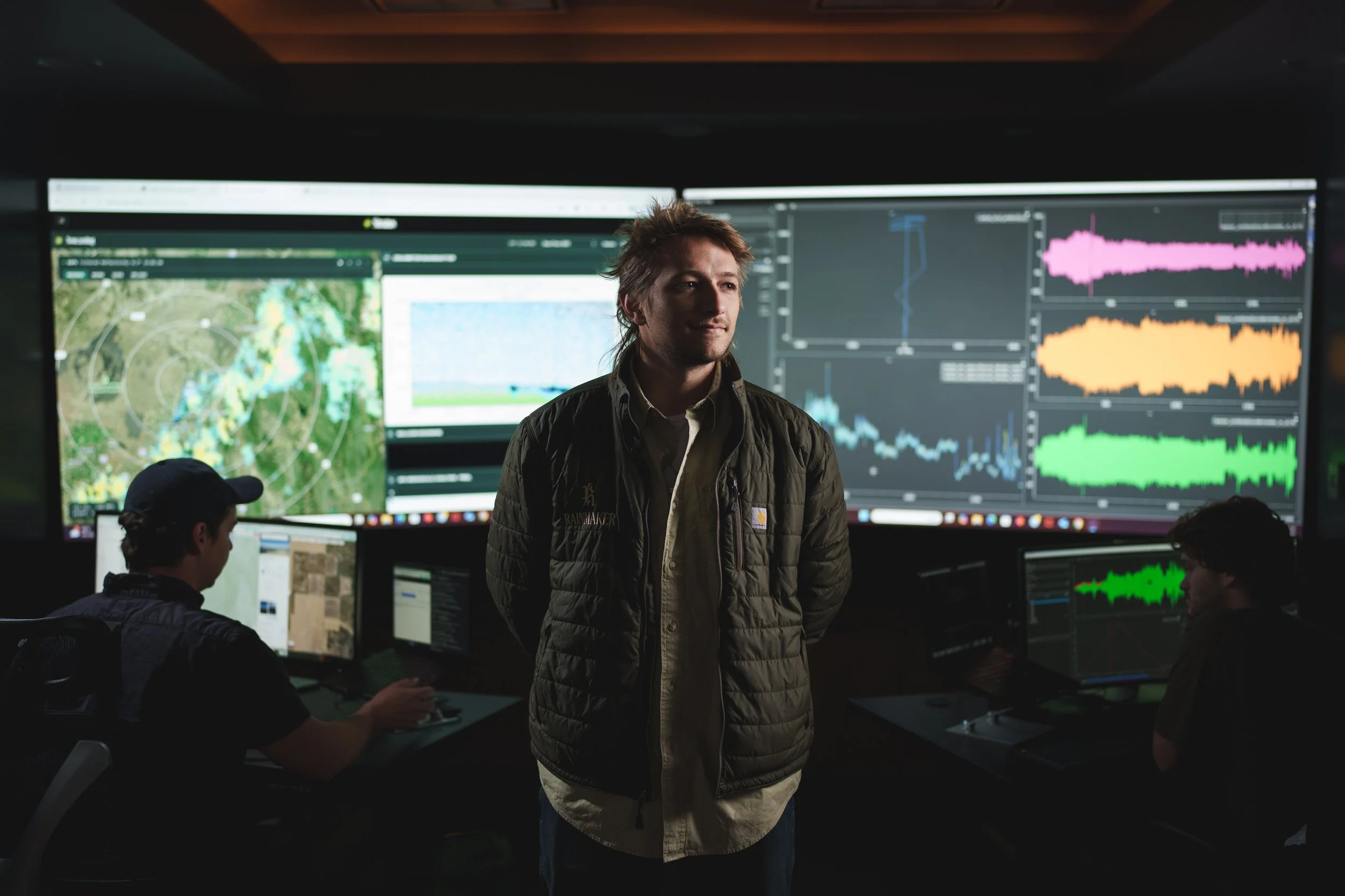 A man stands in front of a large wall of monitors displaying weather maps and graphs, with two other people working on computers beside him in a control room.