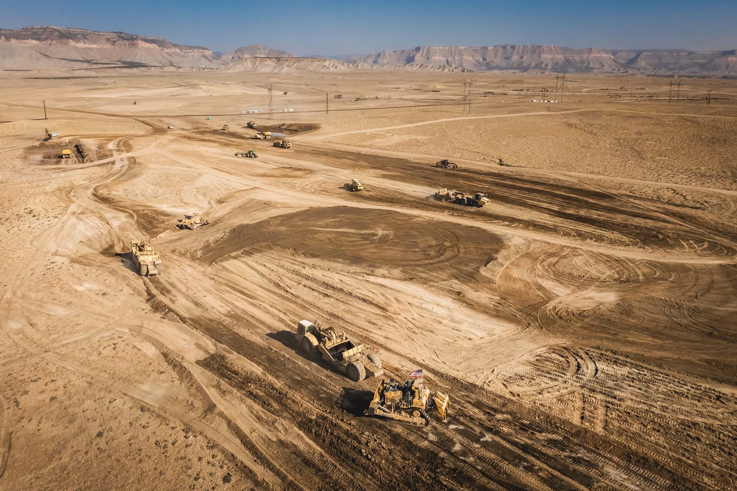 Construction site with multiple bulldozers and earth-moving equipment working on a large, flat desert area with distant mountains under a clear sky.