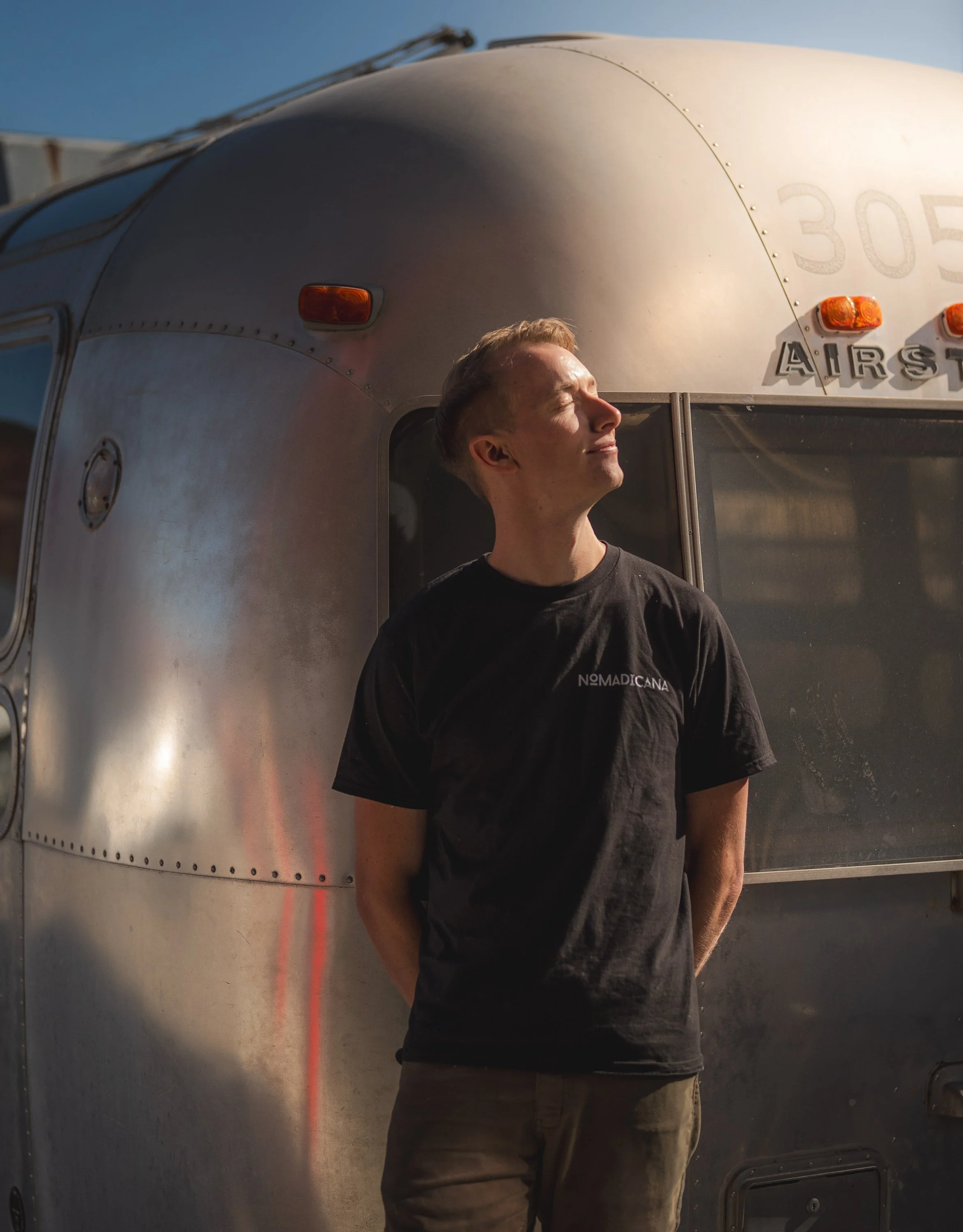 A young man stands with his eyes closed and face tilted upward, basking in sunlight in front of a silver Airstream trailer.