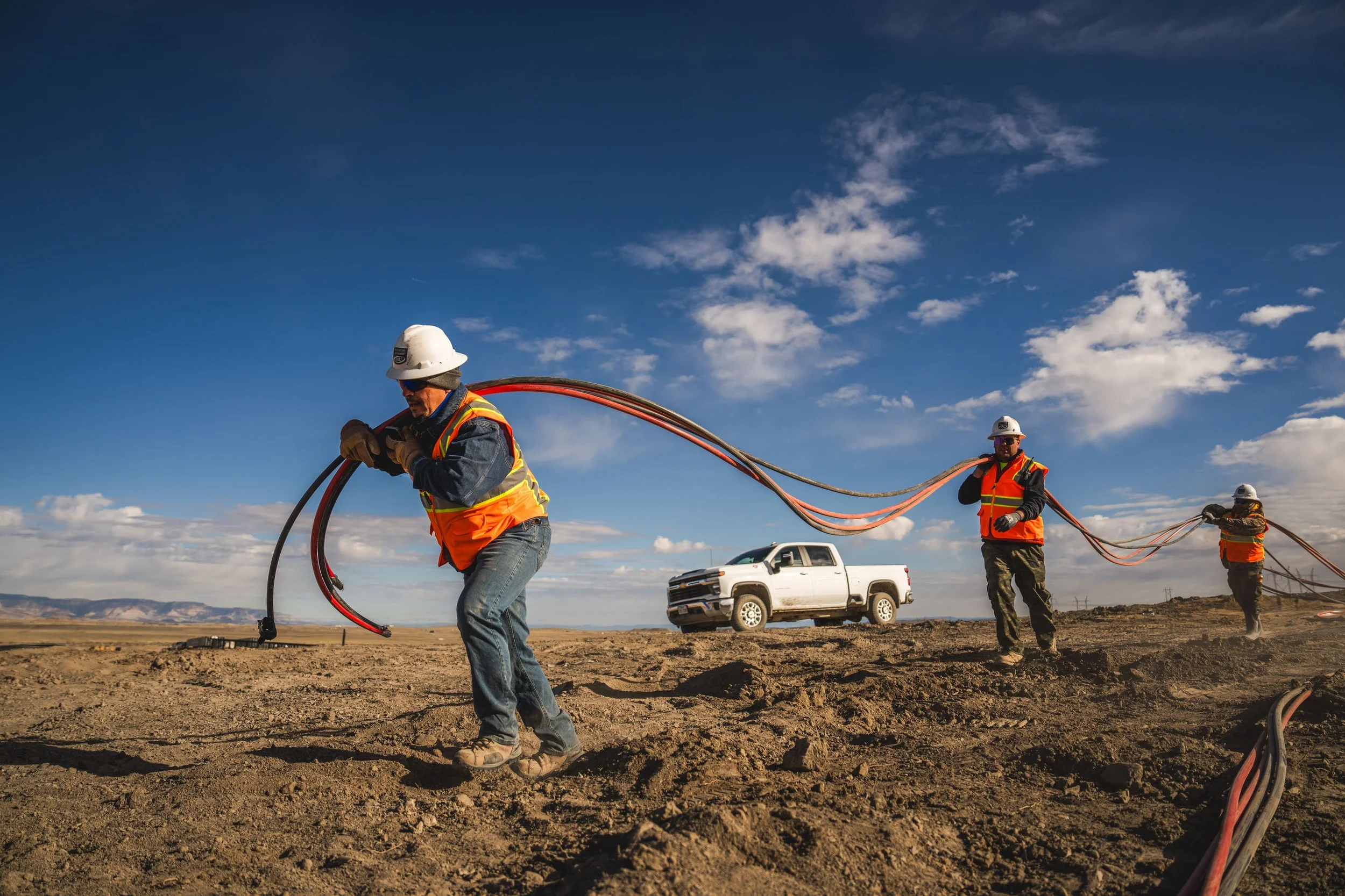 Workers in safety vests and helmets laying electrical cables on a dirt field with a white pickup truck in the background and a blue sky with some clouds.