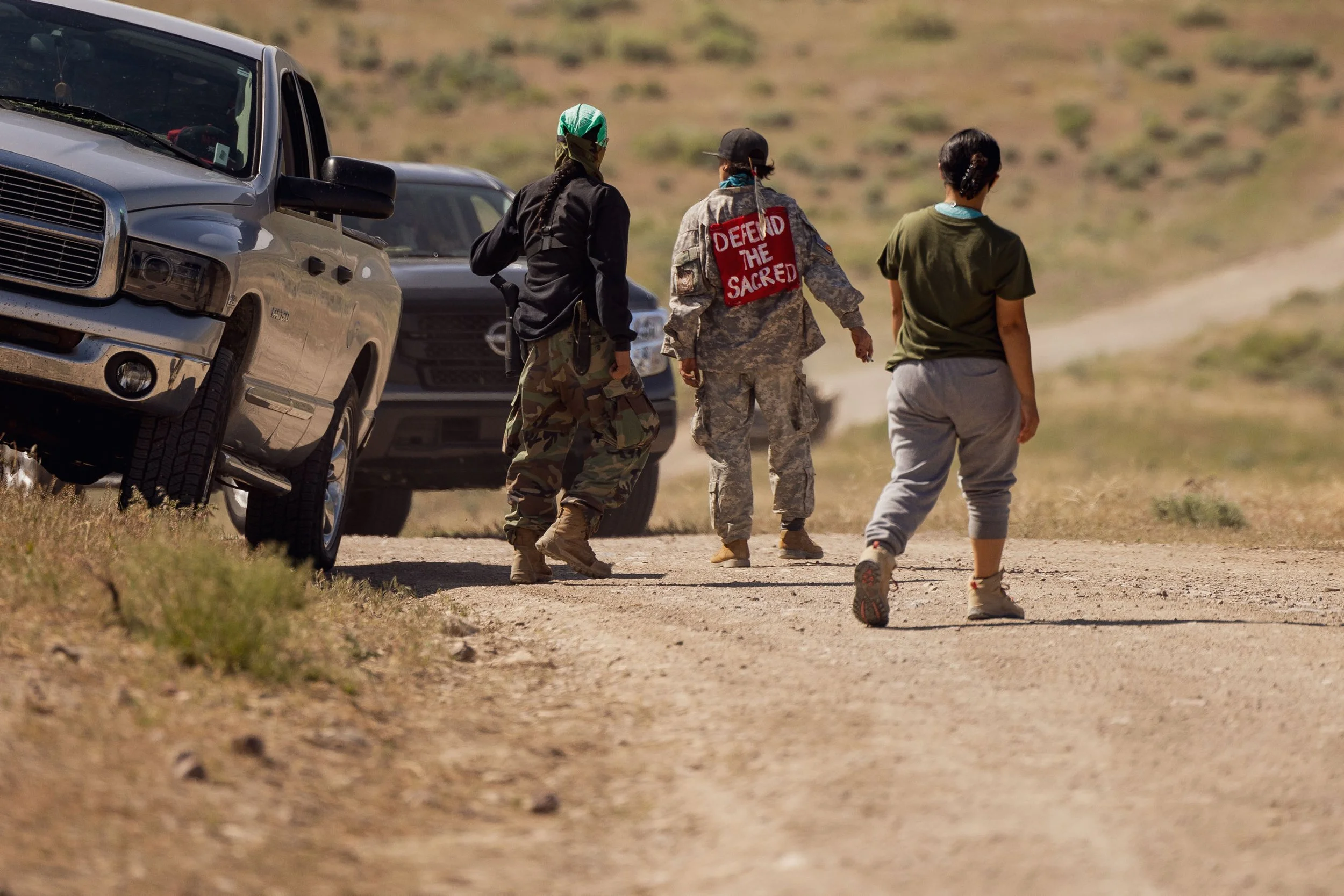 Three women walking on a dirt road in a desert landscape with two parked vehicles nearby.