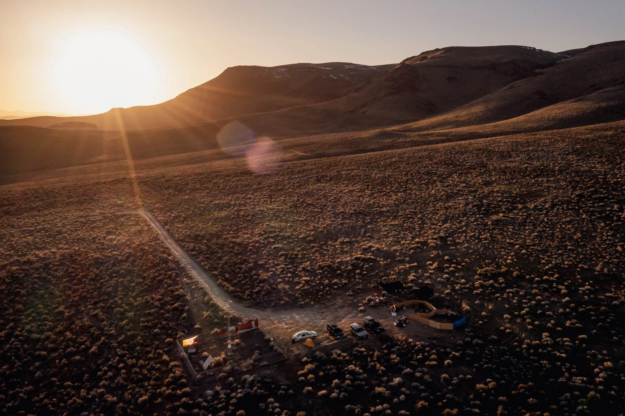 Aerial view of a small campsite with tents and cars at sunset on a hillside covered in bushes.