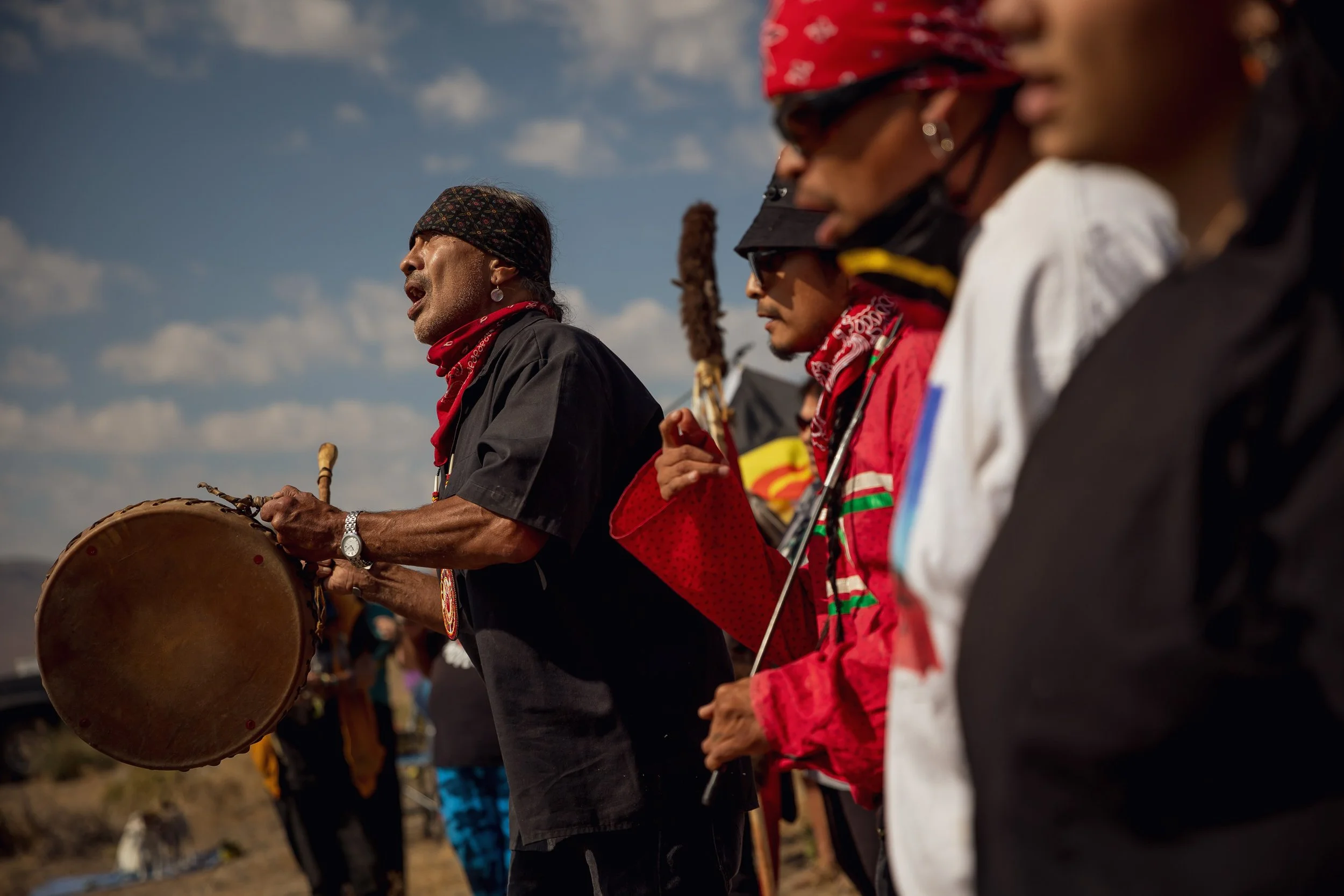 A group of Indigenous people participating in a cultural ceremony outdoors, with one person playing a drum and others holding objects, under a partly cloudy sky.