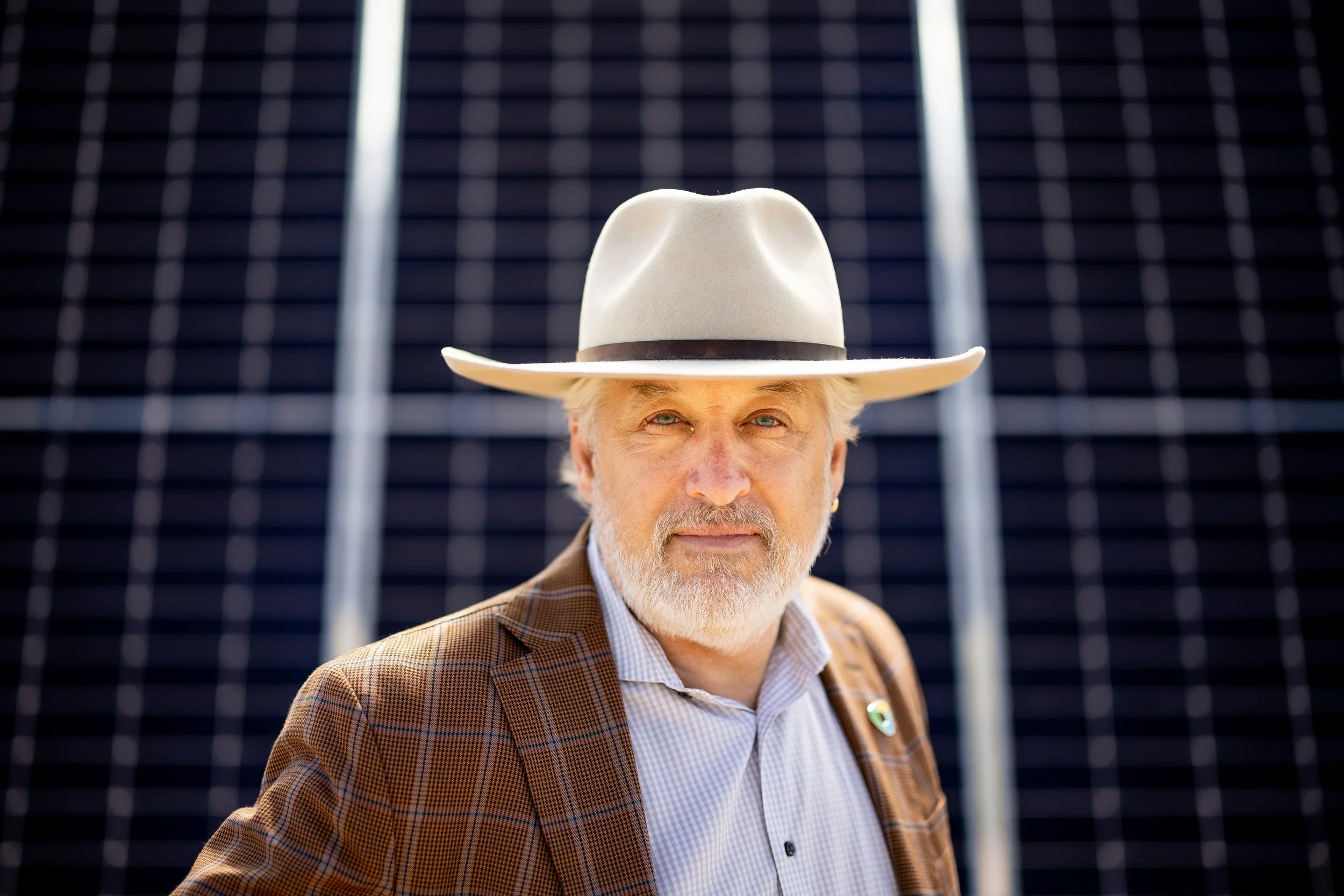 A man with a white beard wearing a beige cowboy hat, a plaid blazer, and a light-colored shirt stands in front of solar panels.