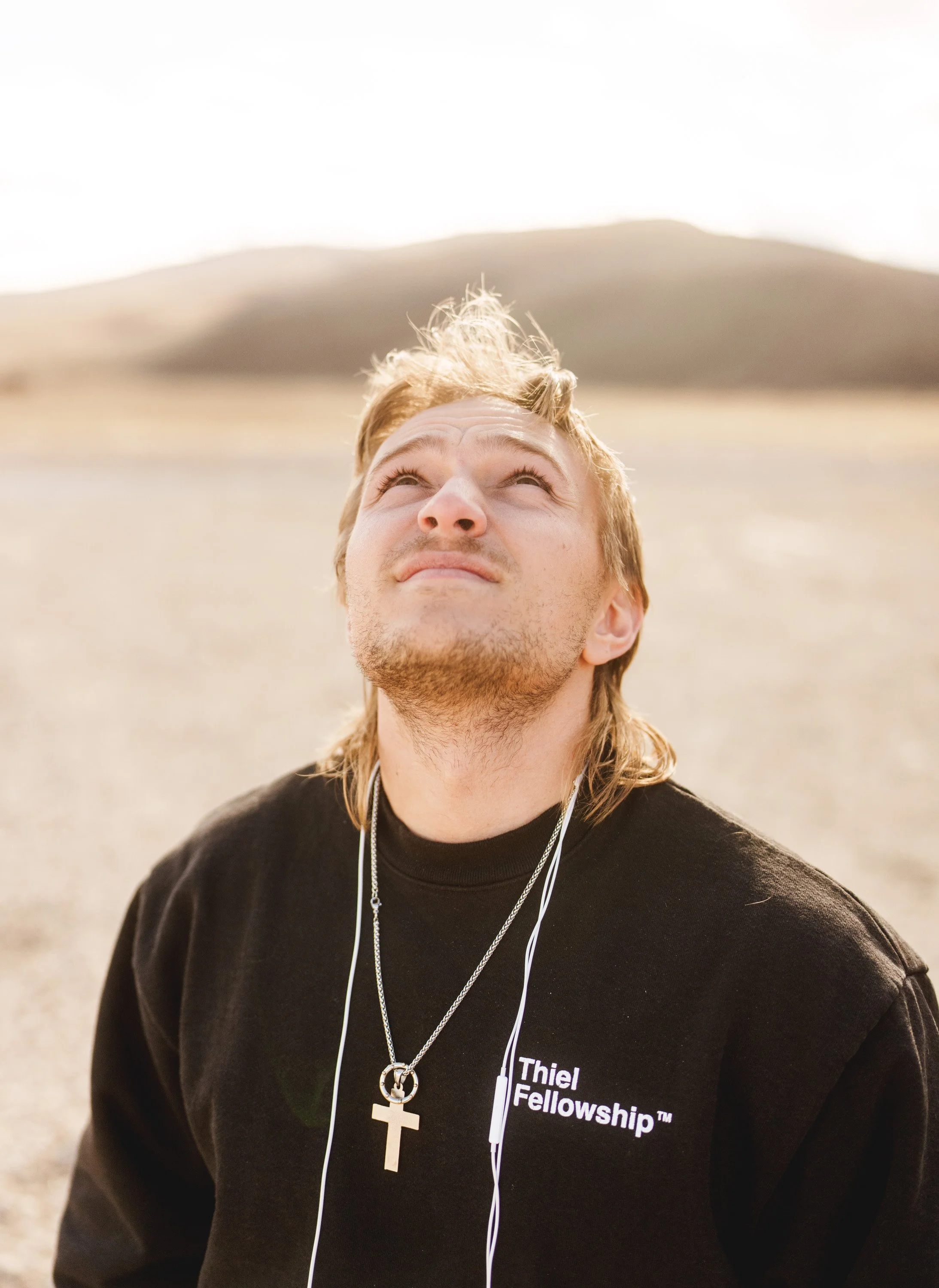 Young man with blond hair gazing upward, wearing a black Thiel Fellowship sweatshirt, a silver cross necklace, and earphones, outdoors during daytime with a blurred landscape background.