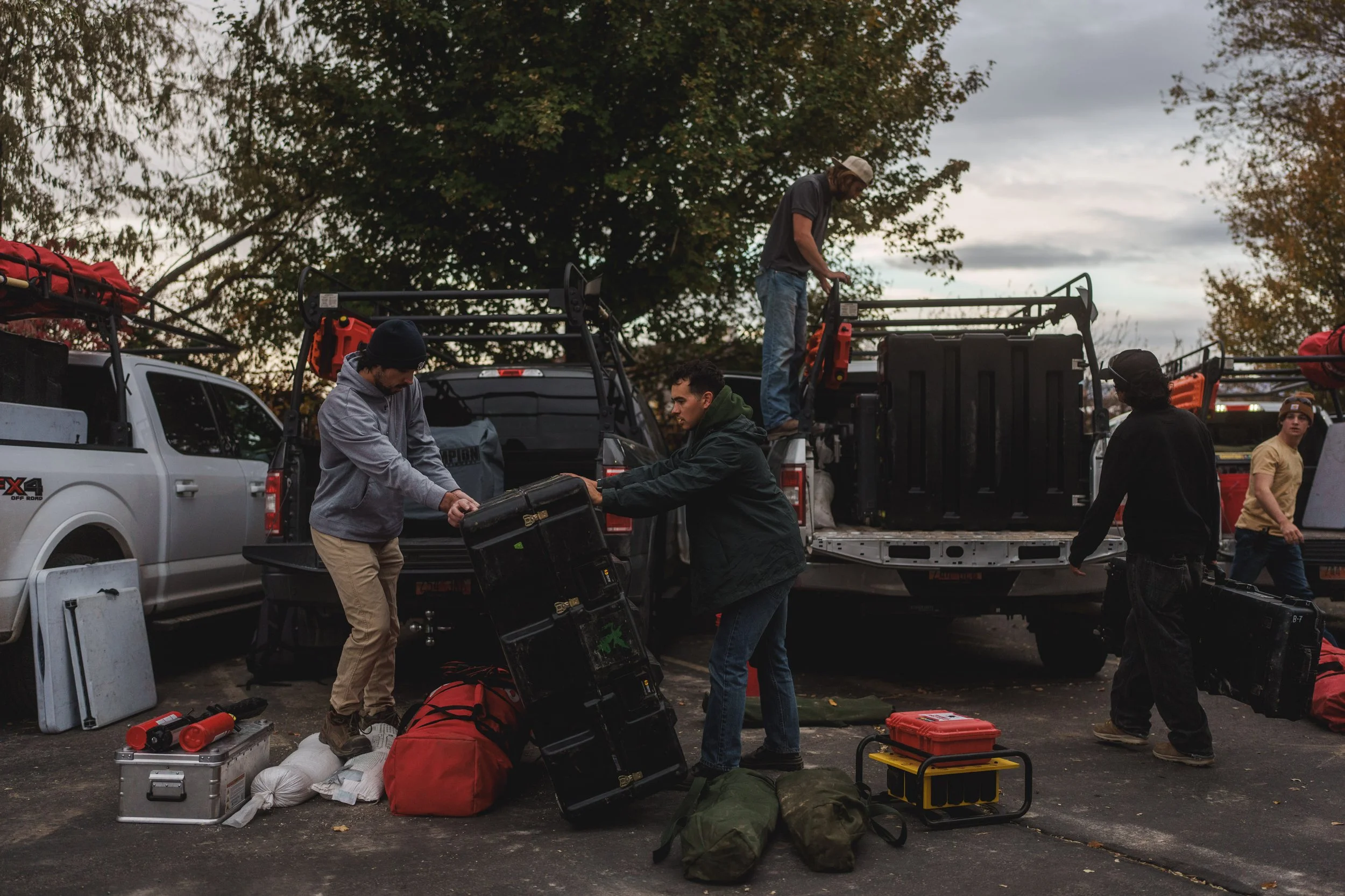 Group of young people preparing camping or outdoor gear beside pickup trucks during late afternoon or evening with cloudy sky.