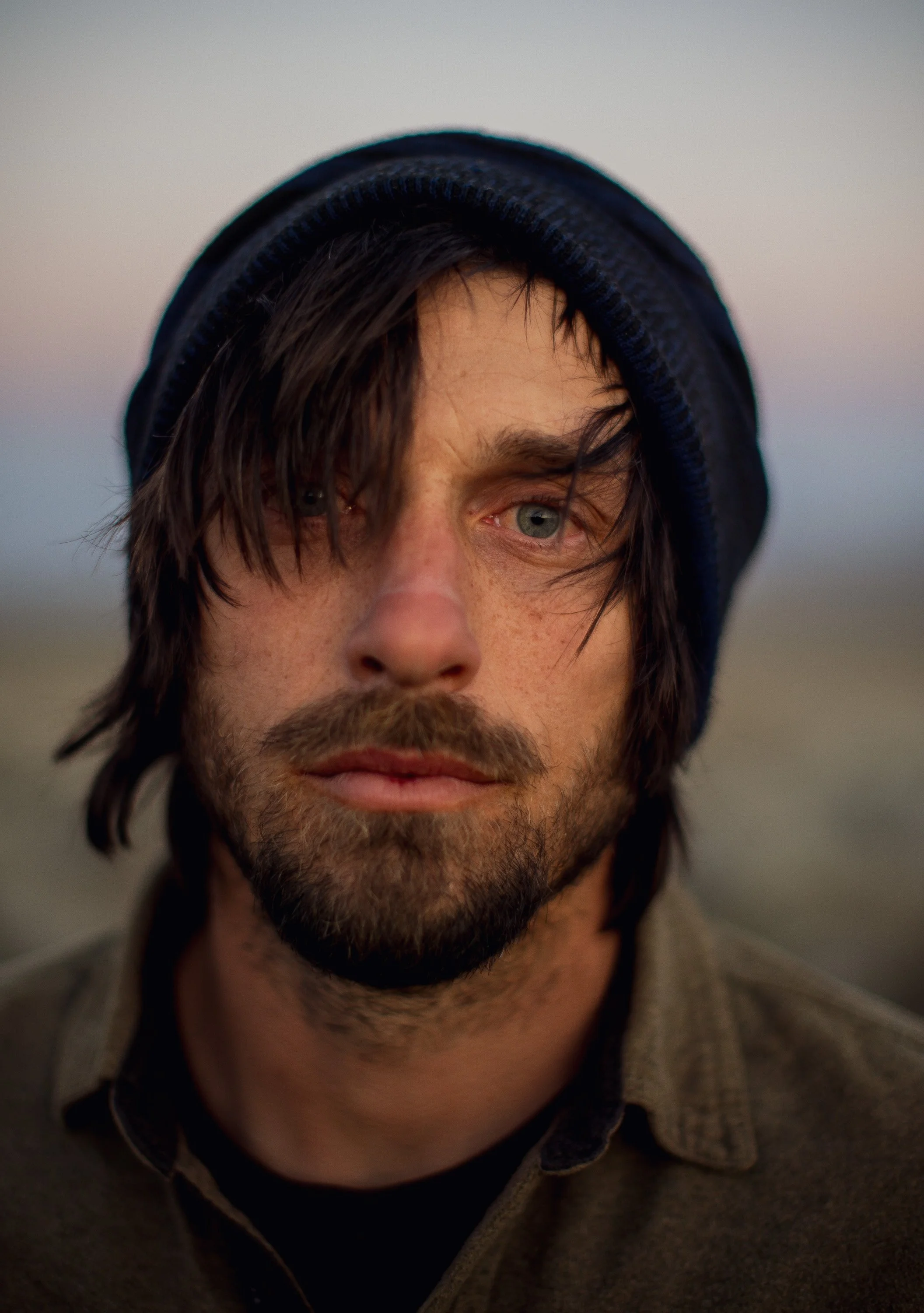 Close-up of a man with shaggy brown hair, a beard, wearing a dark blue beanie, looking into the camera with a neutral expression outdoors at dusk.