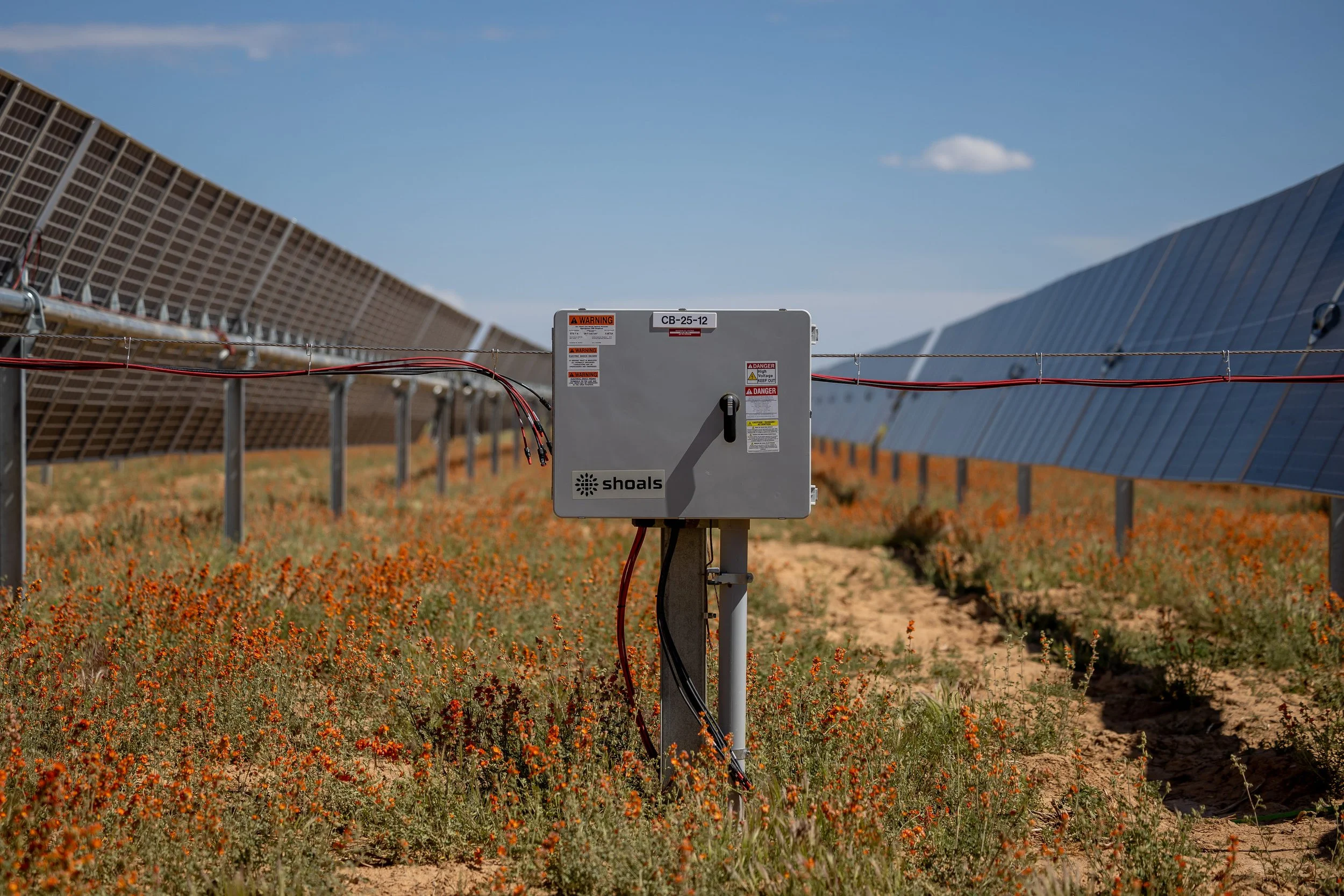 Solar panels in a field with electrical equipment in the foreground under a clear blue sky.
