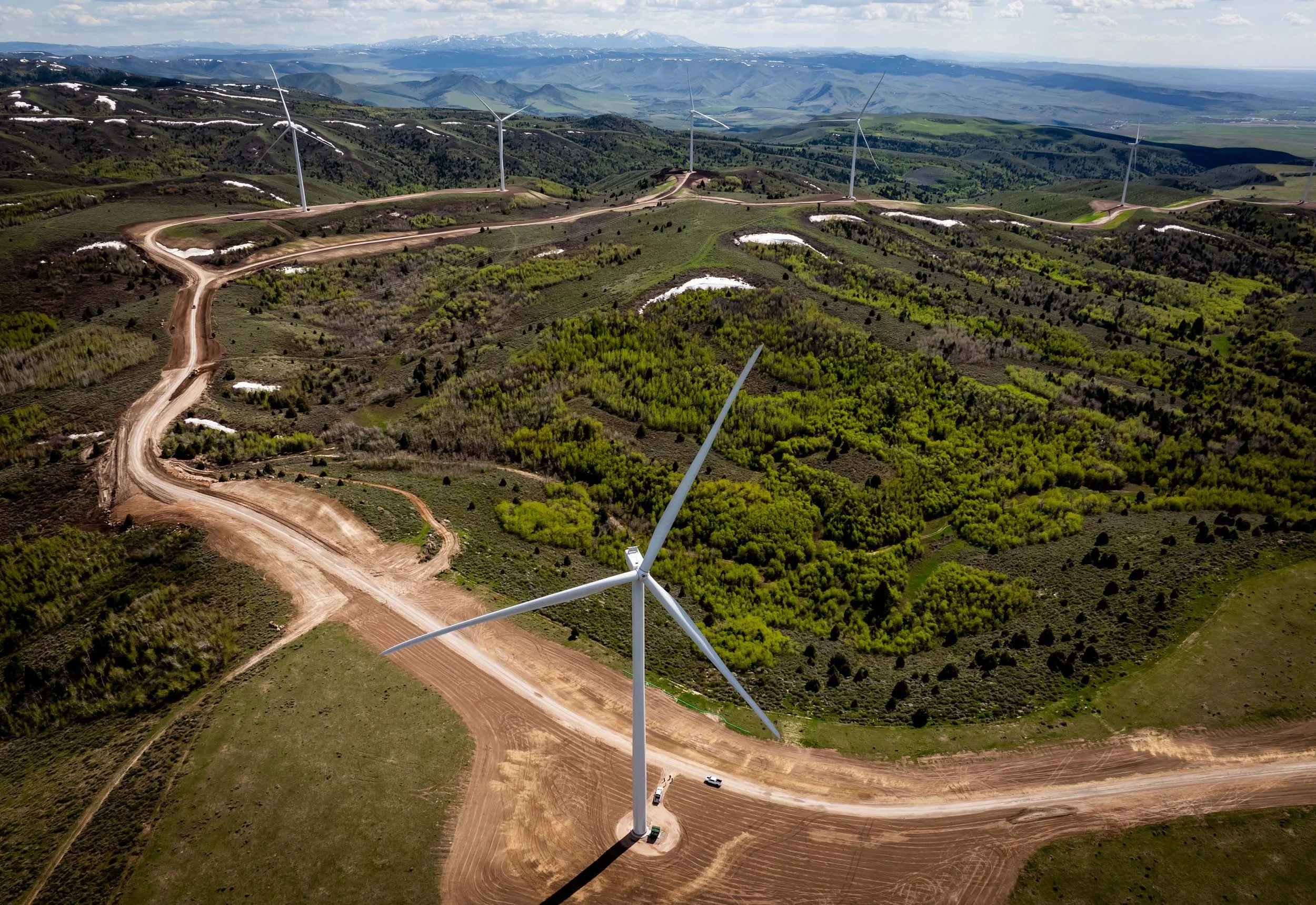 Aerial view of a wind farm with several wind turbines on a green landscape with dirt roads, rolling hills, and patches of snow in the background.