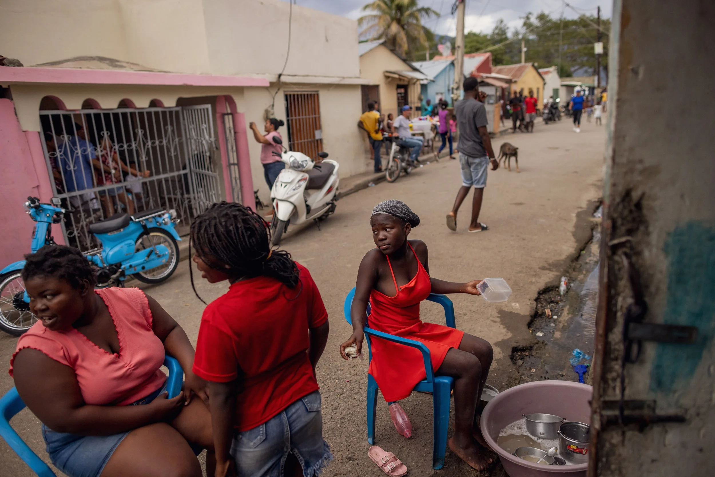 Two women seated on blue chairs and a girl in a red dress sitting on a blue chair on a street in a neighborhood. The woman on the left wears a pink top and shorts, the woman in the middle wears a red top and denim shorts, and the girl wears a red dress with a grey head covering. The girl holds a plastic food container and sits next to a basin with pots and utensils. People walk and bike on the street, with houses and motorcycles visible in the background.