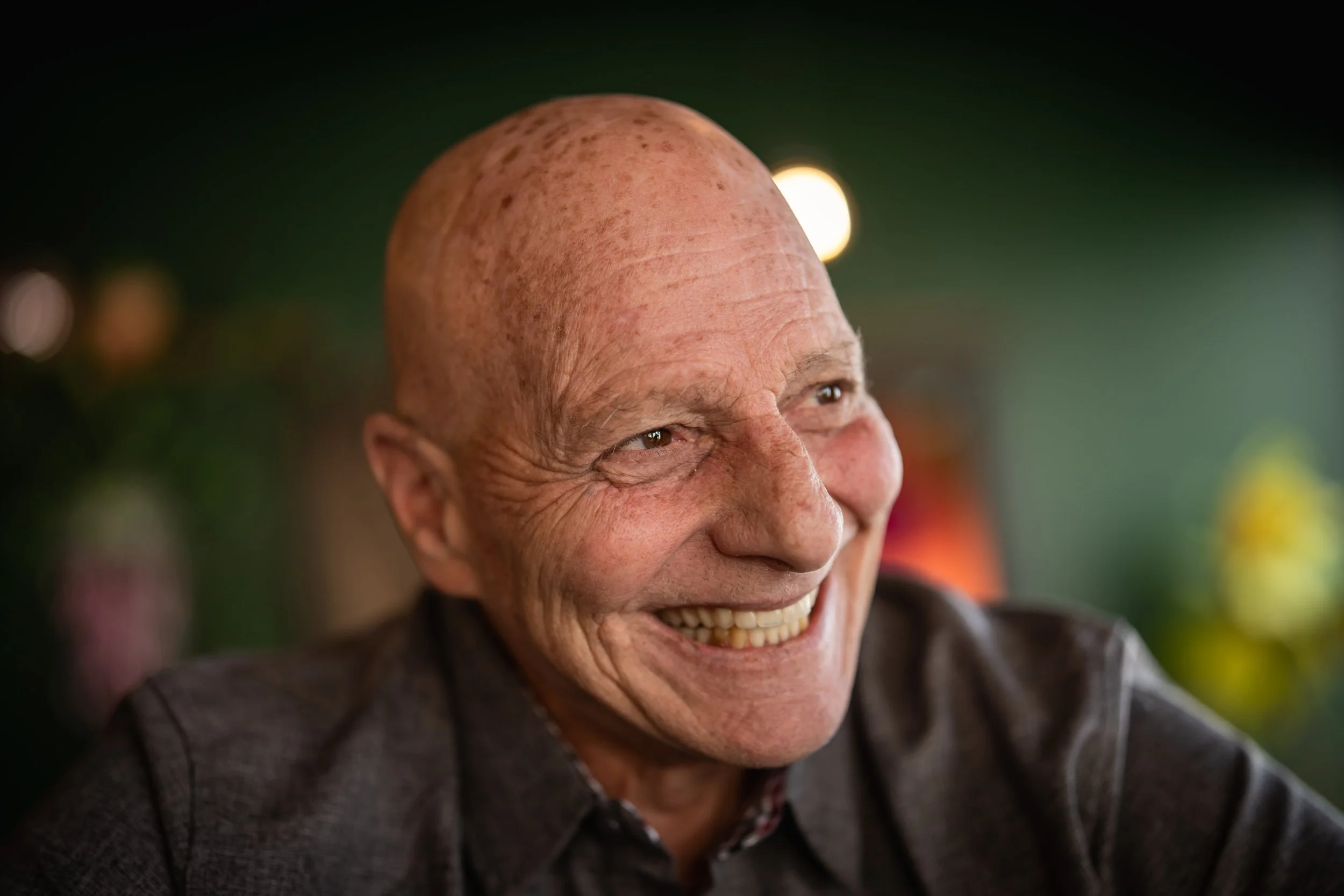 Close-up of smiling elderly man with a bald head, wrinkles, and wearing a dark blazer, in a warmly lit setting.