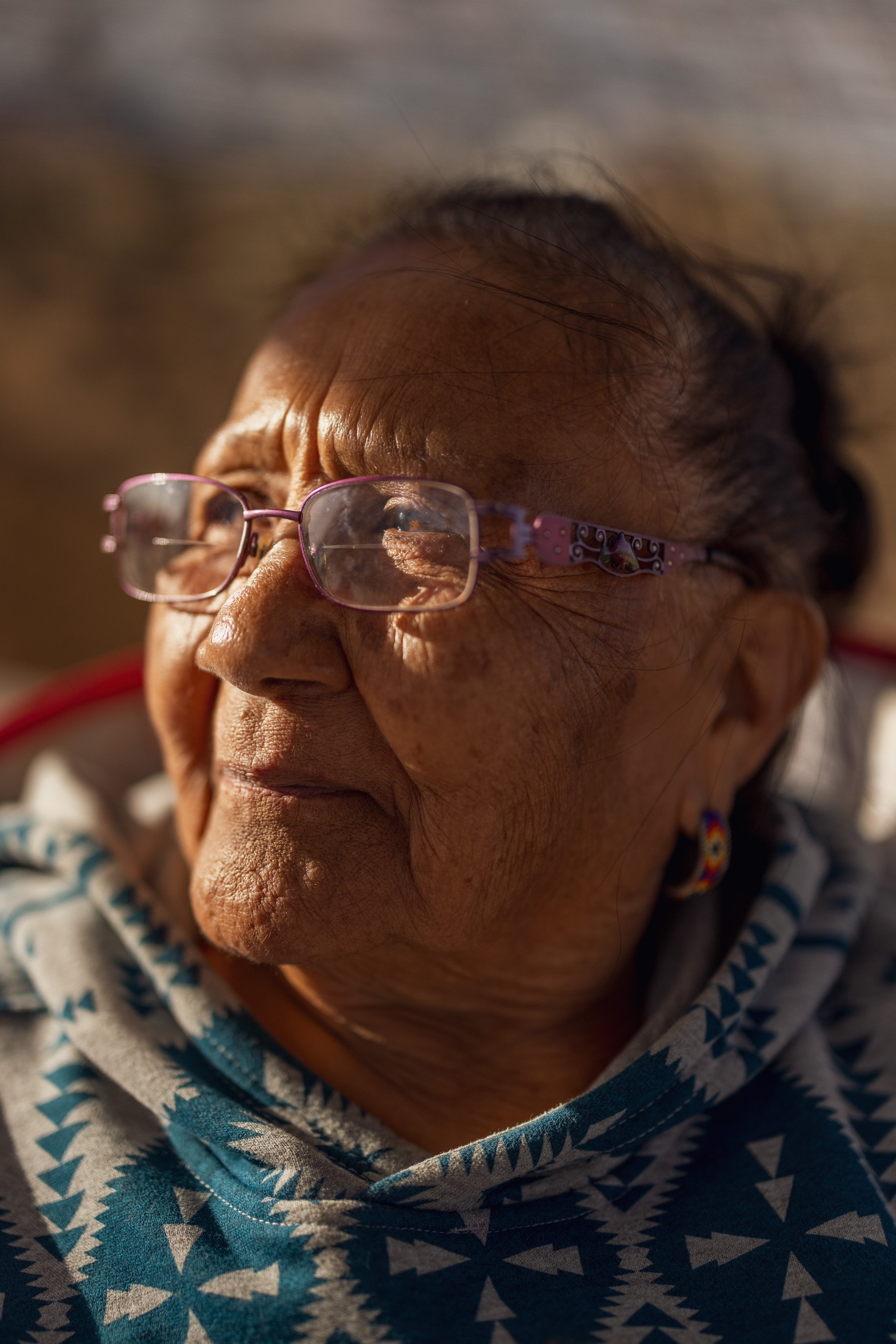 Close-up of an elderly woman with glasses, wearing a blue patterned hoodie, and a colorful earring, looking contemplatively into the distance.