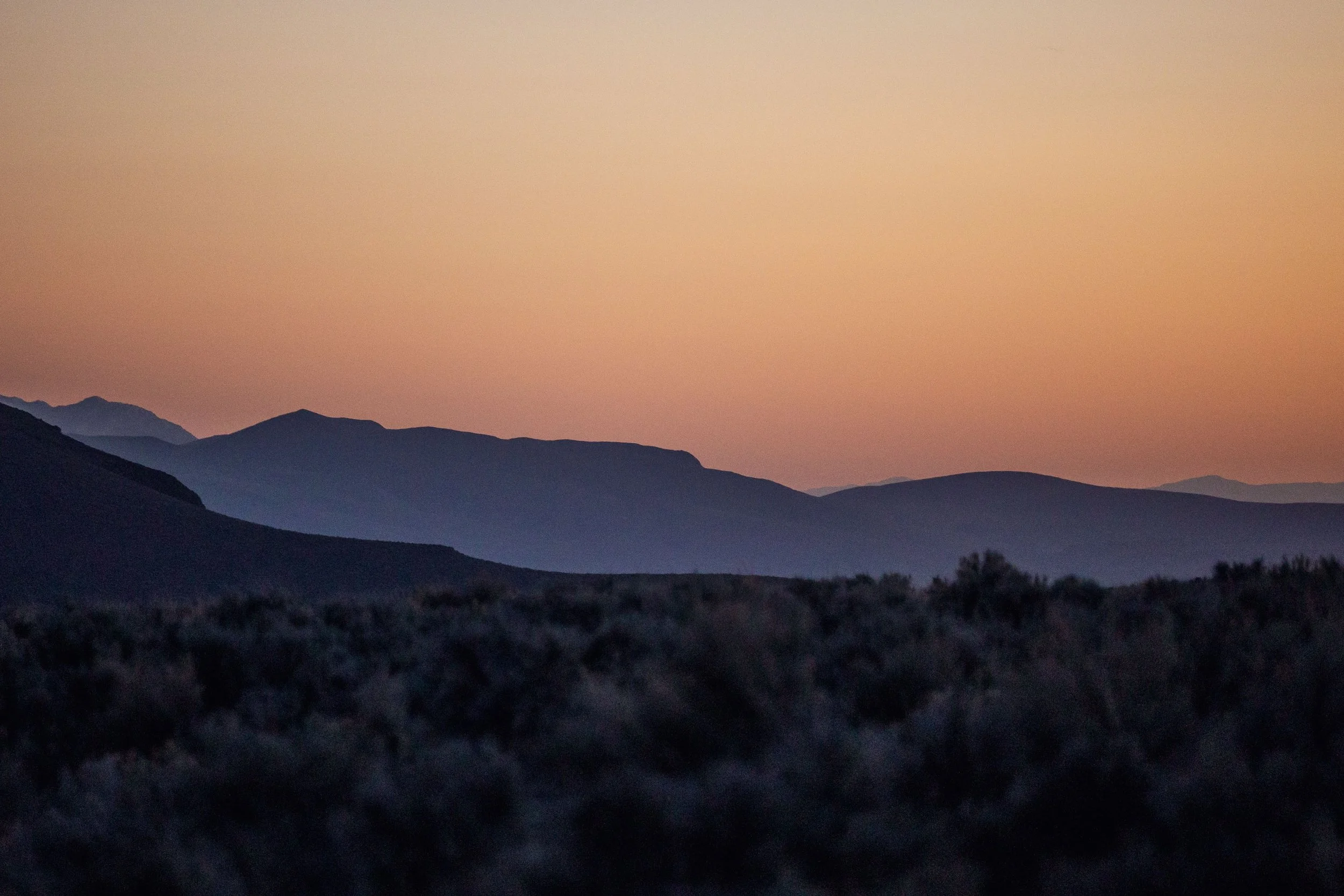 A landscape of mountains at sunset, with a pink-orange sky and dark silhouettes of distant hills and foreground shrubs.