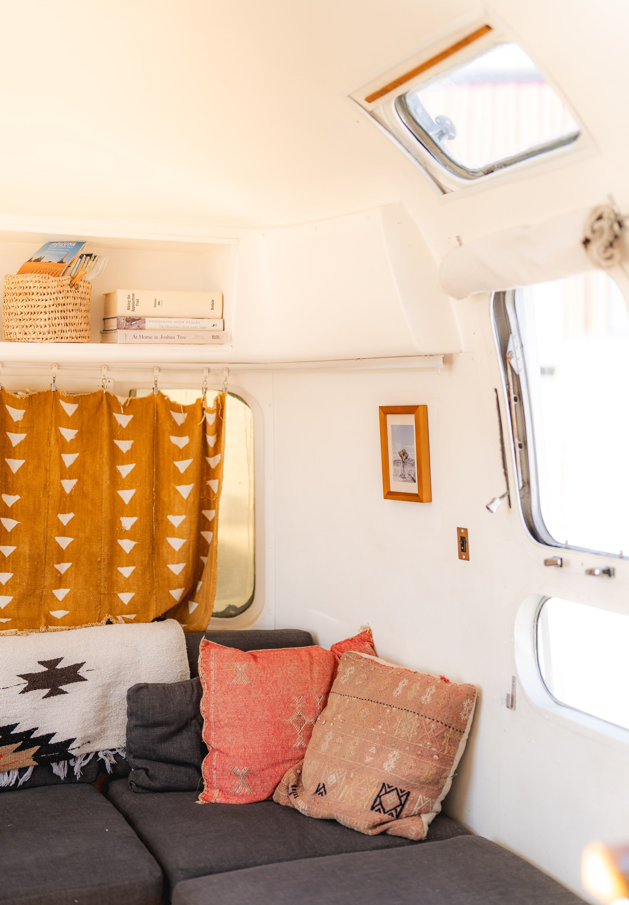 Interior of a cozy living space with a dark sofa adorned with decorative cushions, a window with orange patterned curtains, a bookshelf with books and a woven basket, a small framed picture, and irregularly shaped windows on a white wall.
