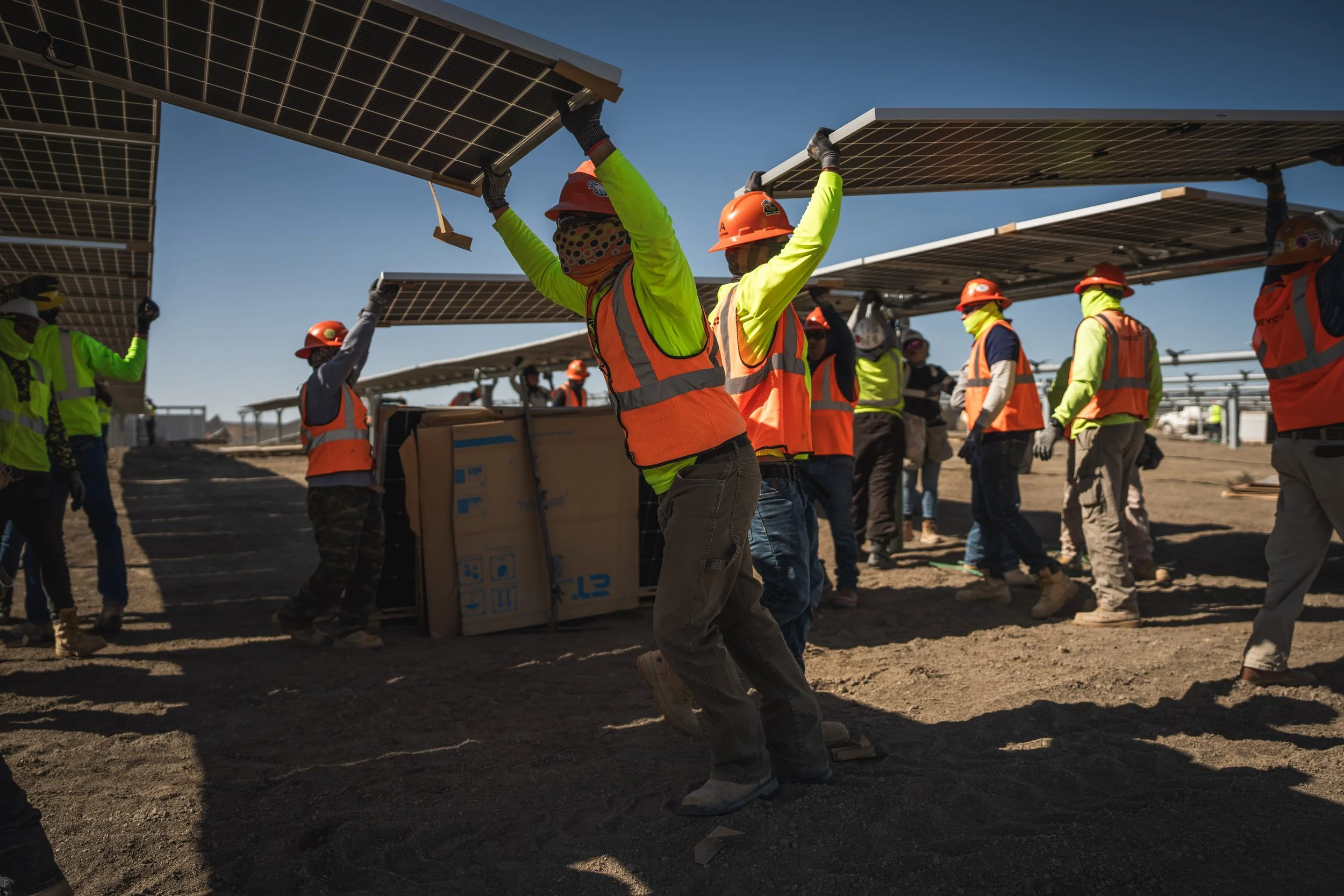Group of workers installing solar panels outdoors, wearing safety gear including helmets, vests, and gloves, handling solar panel modules under a clear blue sky.