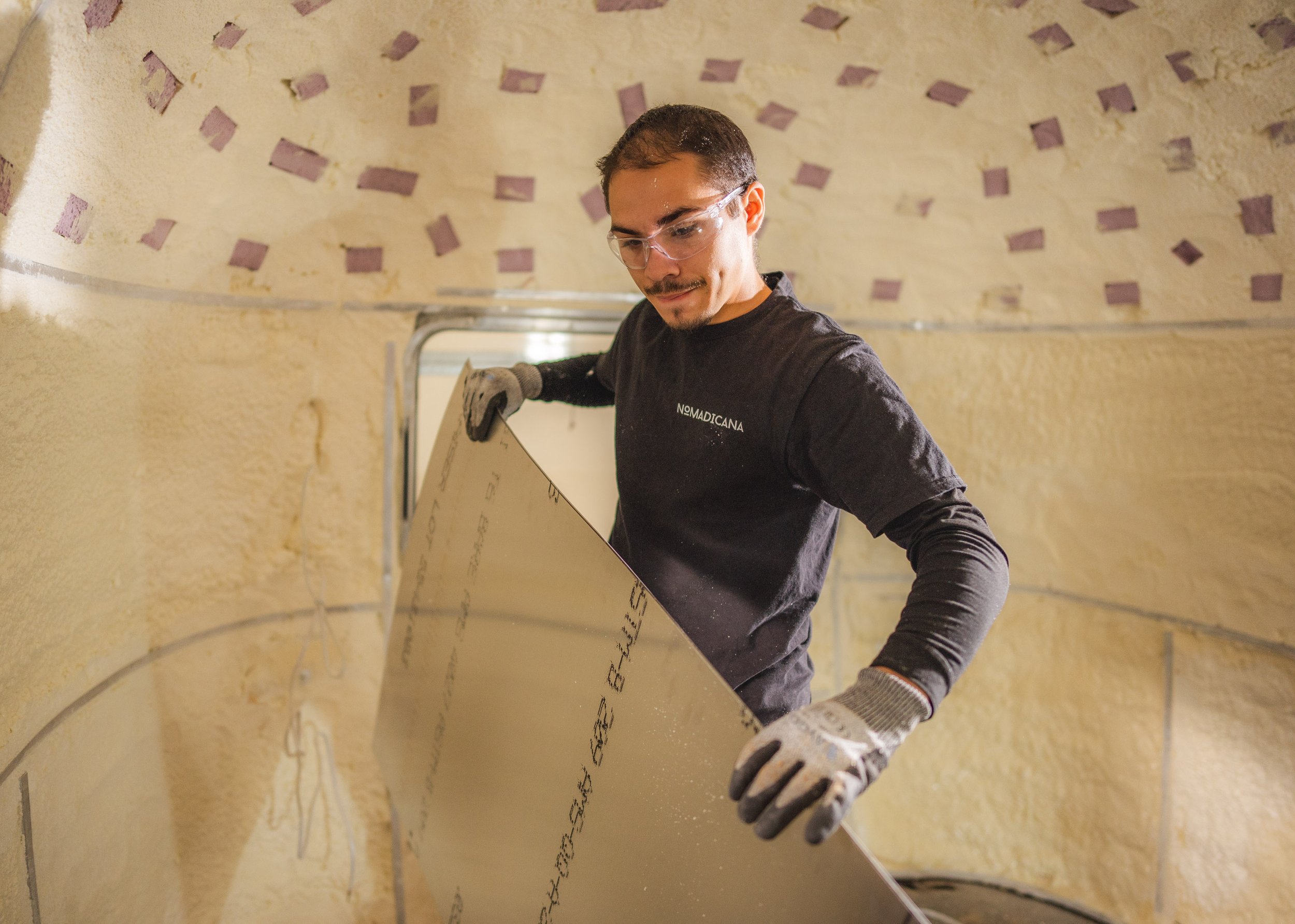 A man wearing safety glasses, a black shirt, and work gloves installs a sheet of drywall inside a textured yellow-walled room.