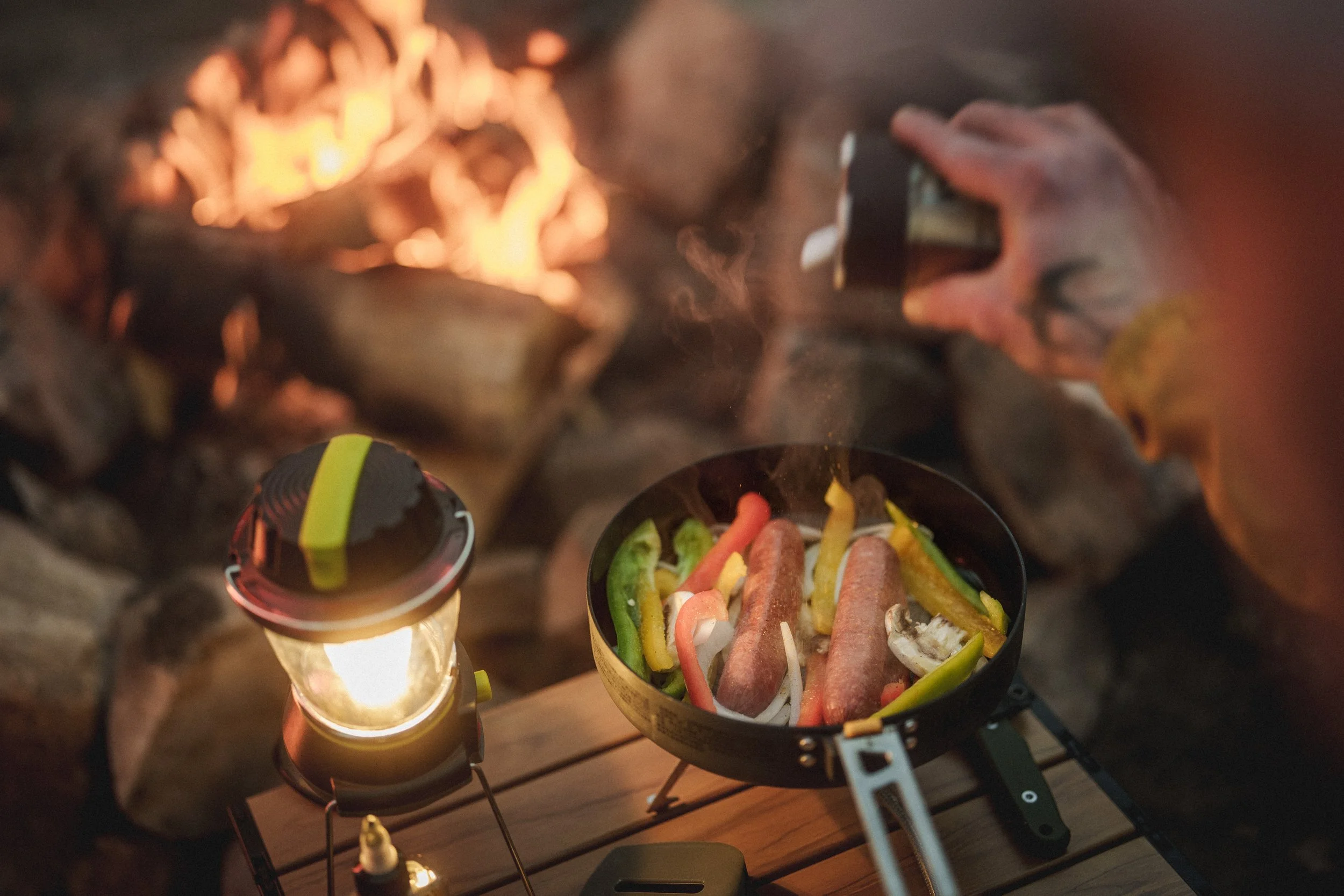 Person cooking hot dogs and vegetables over a campfire on a portable stove with a lantern nearby.