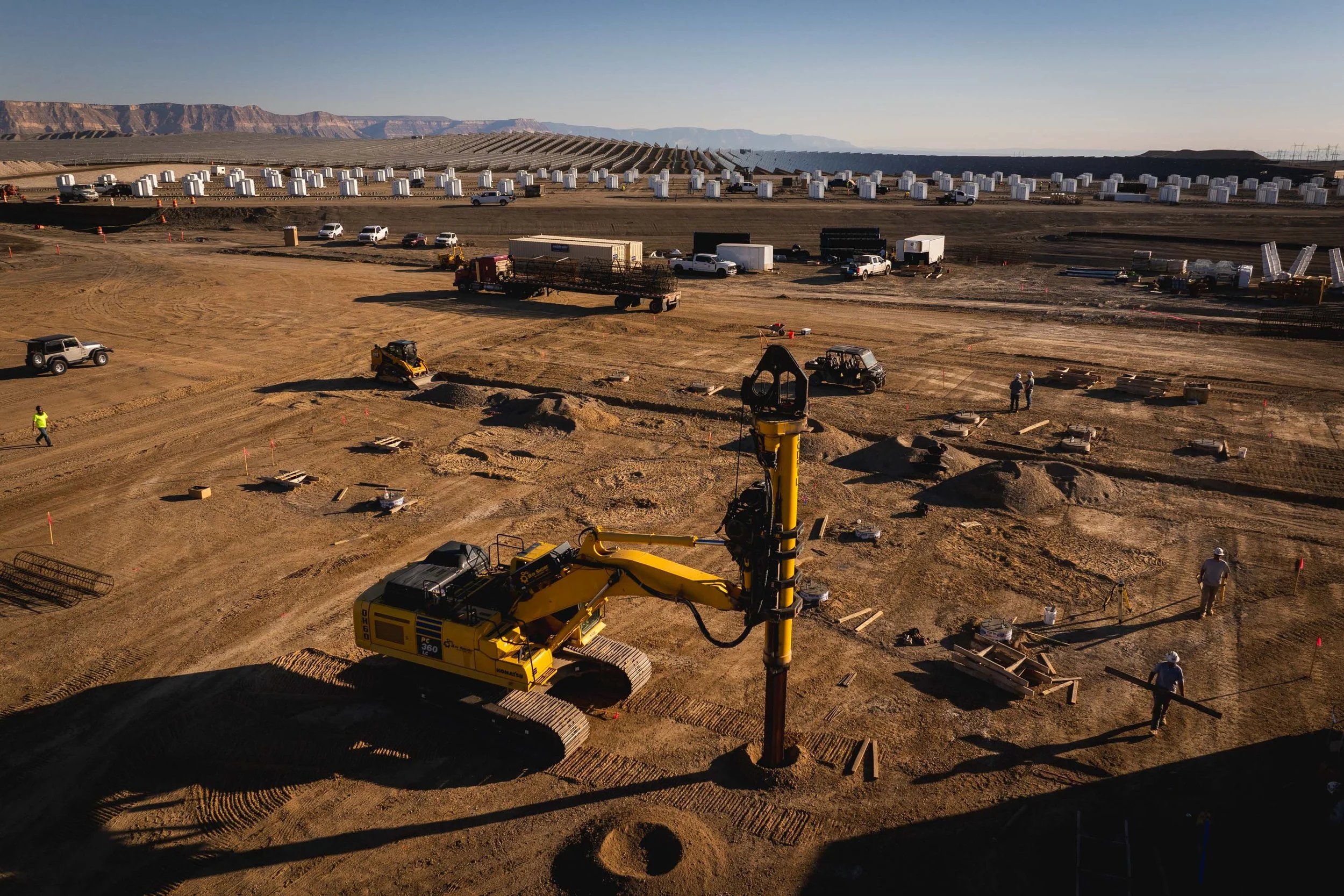 Construction site with a large yellow crane, multiple workers, and equipment, with solar panels and mountains in the background.