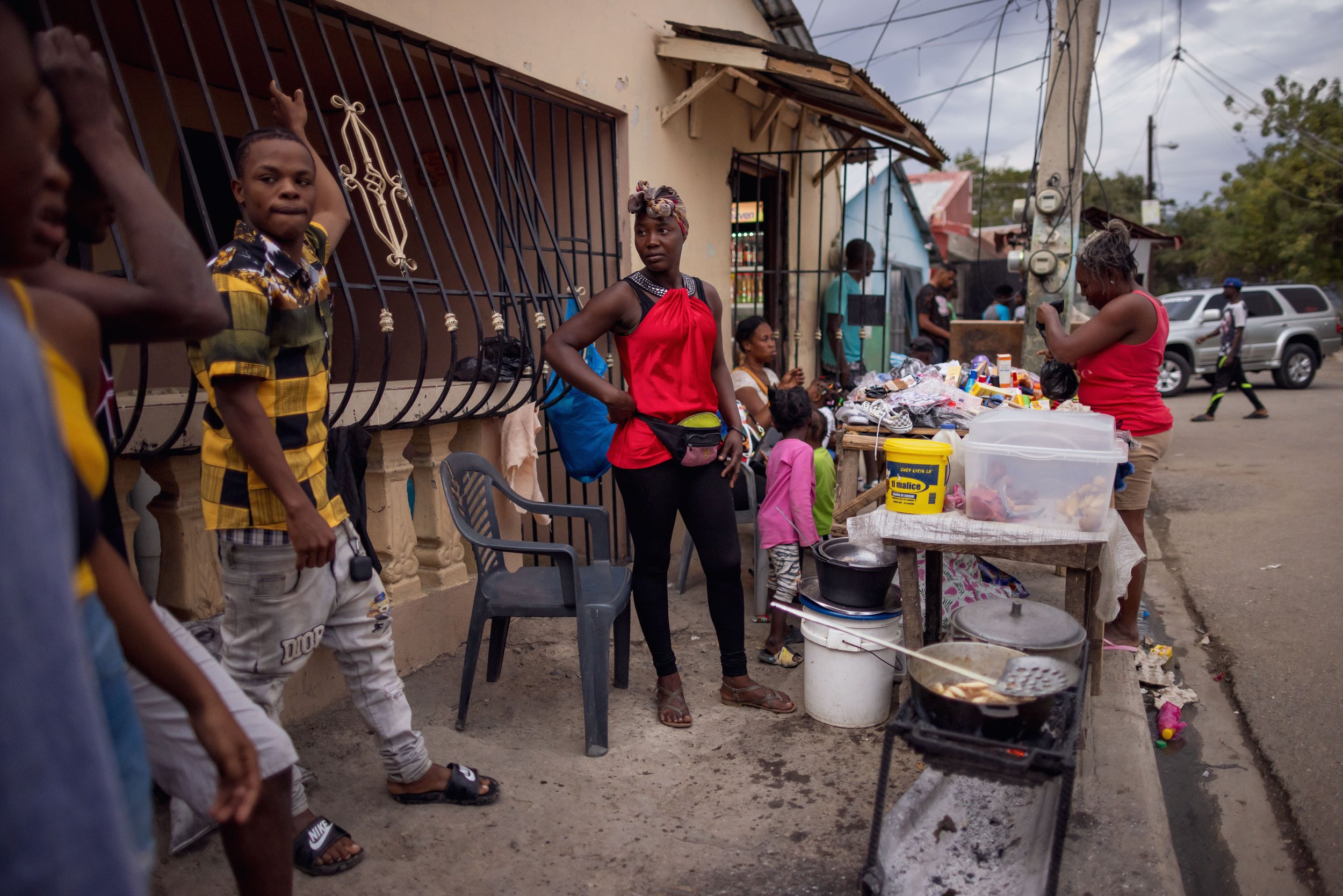 Street scene showing people gathered around a small food stall with various items, a woman in a red top standing with hands on hips, children and adults nearby, parked cars in the background, and an overcast sky.