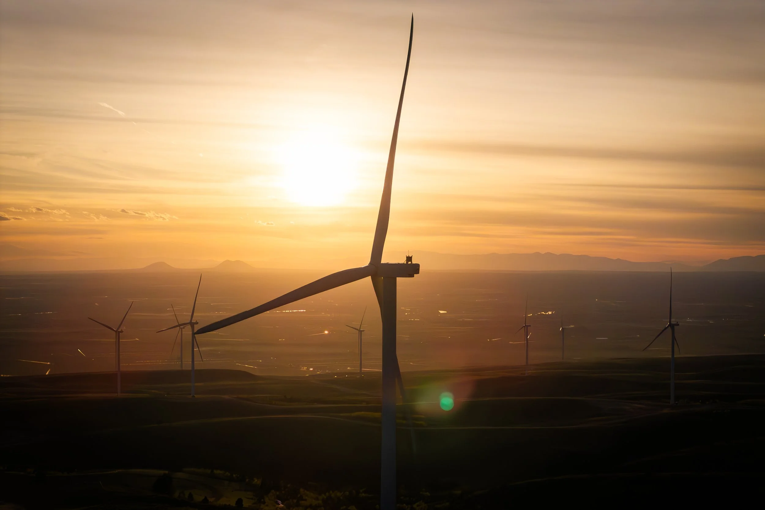 Multiple wind turbines on a hilltop at sunset with a cloudy sky and mountains in the distance.