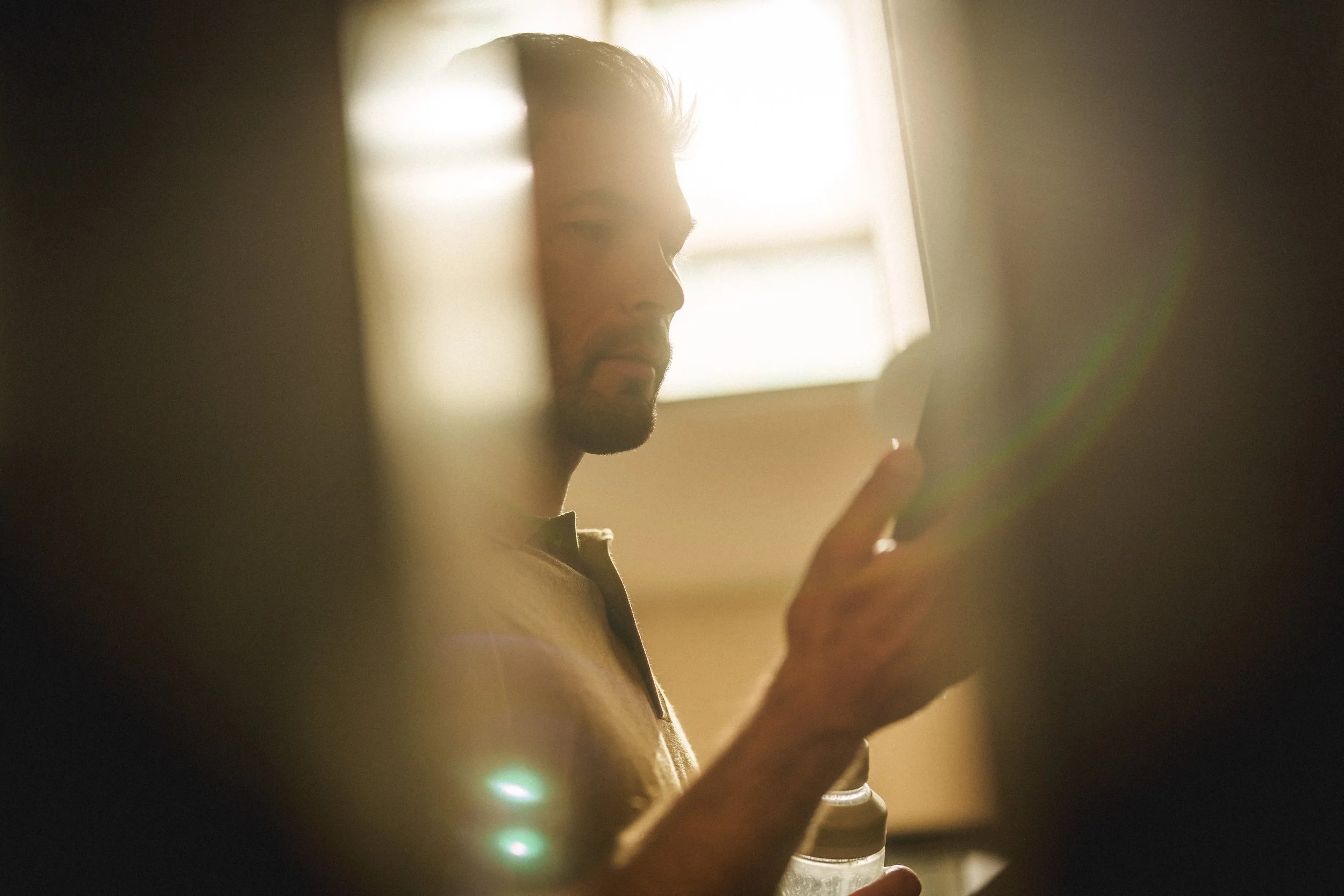 A man with dark hair and a beard looking at his phone while holding a water bottle, with sunlight coming through a window behind him.