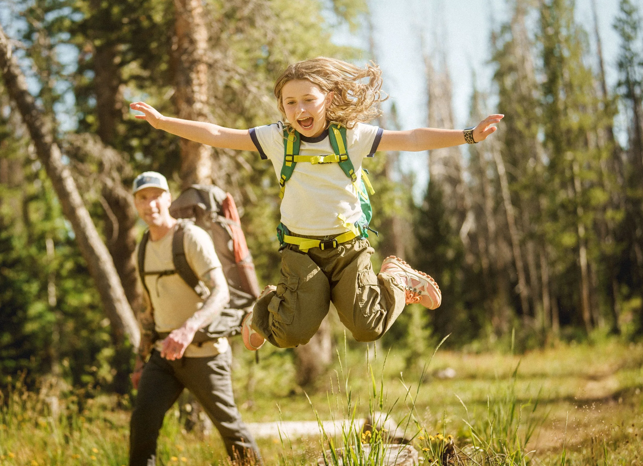 A young girl jumping excitedly in a forest during a hike, with a woman in the background carrying a backpack and wearing outdoor gear, both surrounded by trees and greenery.