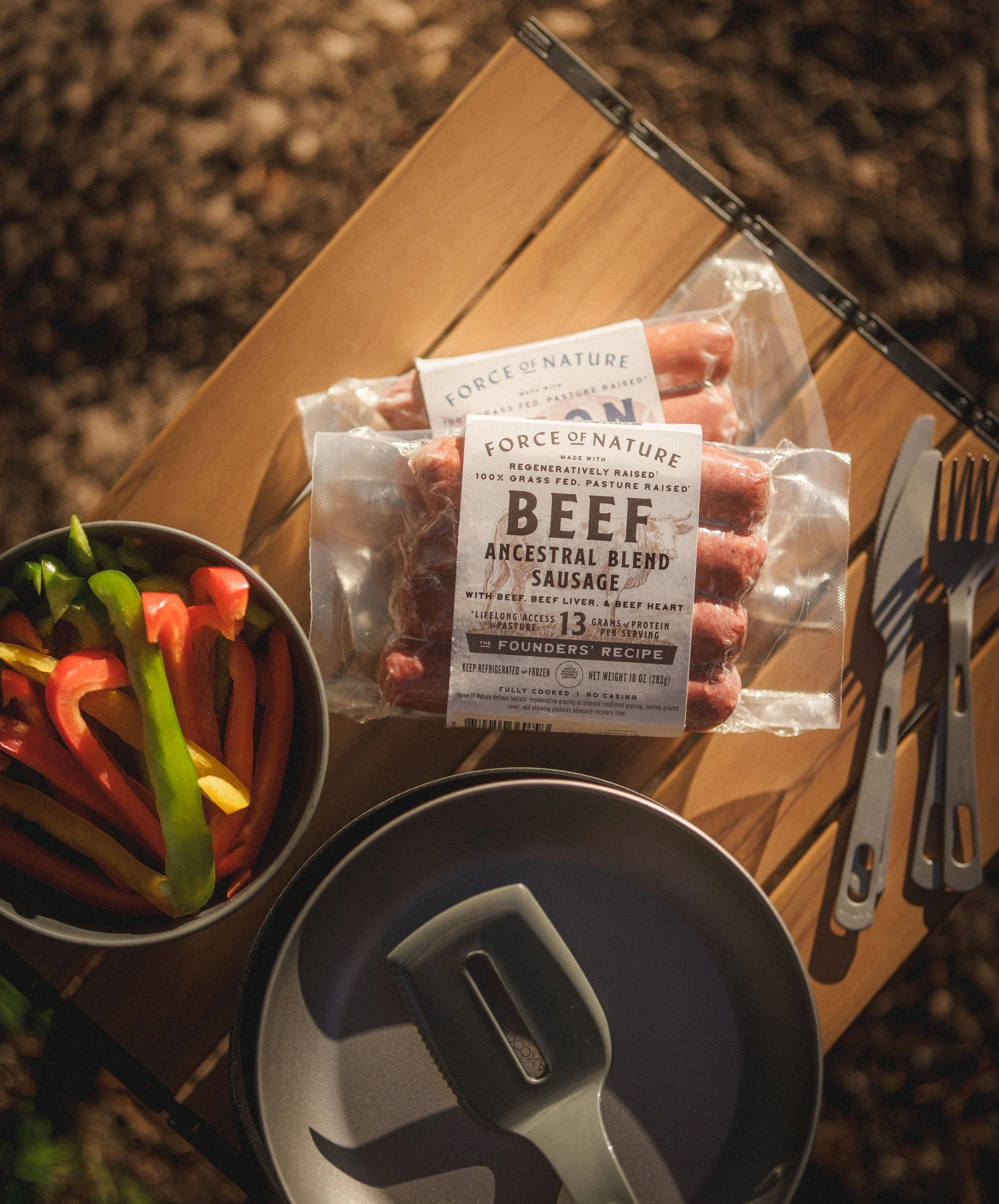 Packaged beef sausage, a bowl of sliced colorful bell peppers, and outdoor utensils on a wooden table.