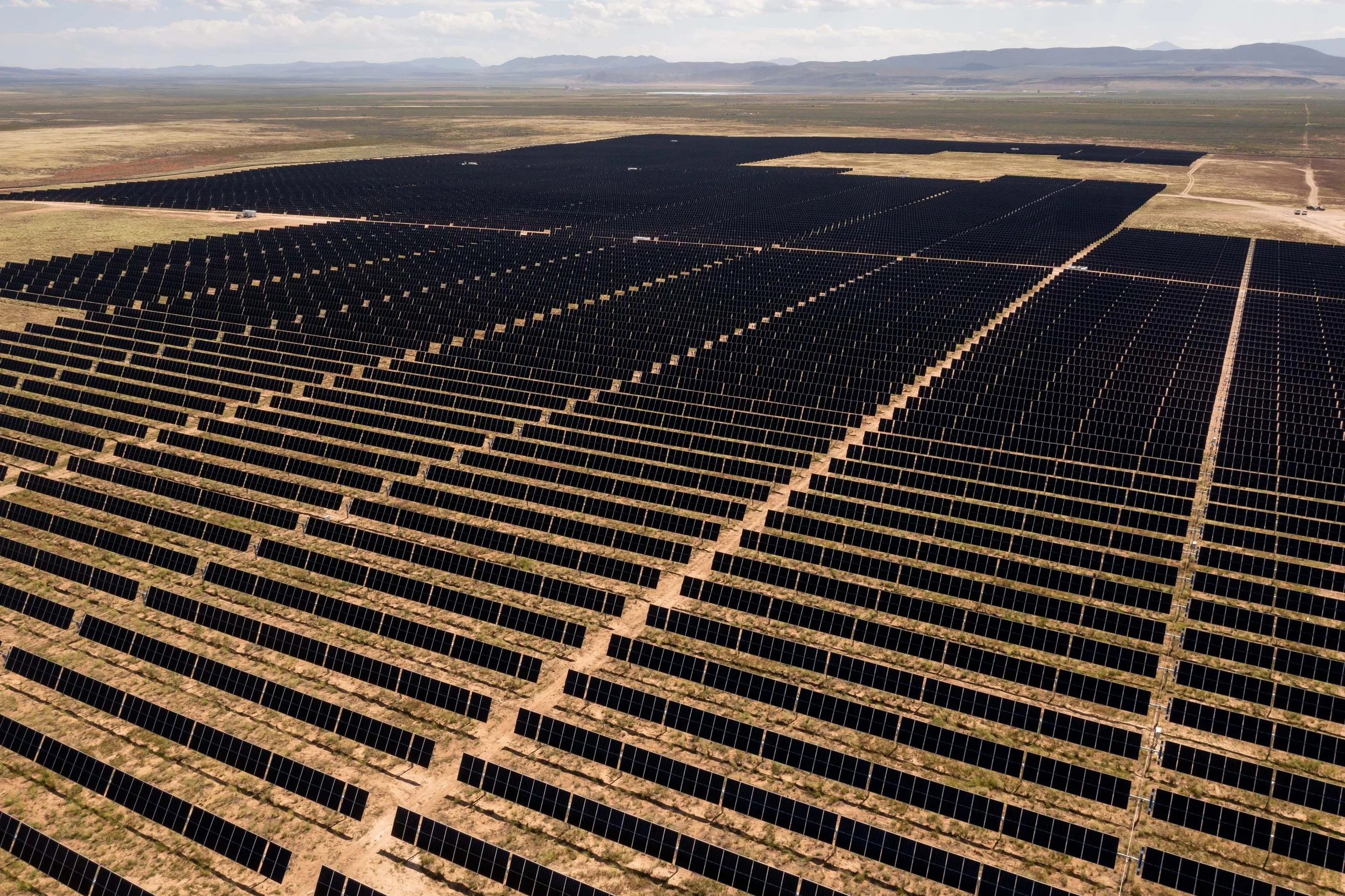A large solar farm with numerous black solar panels arranged in rows on a flat, open landscape with mountains in the background.