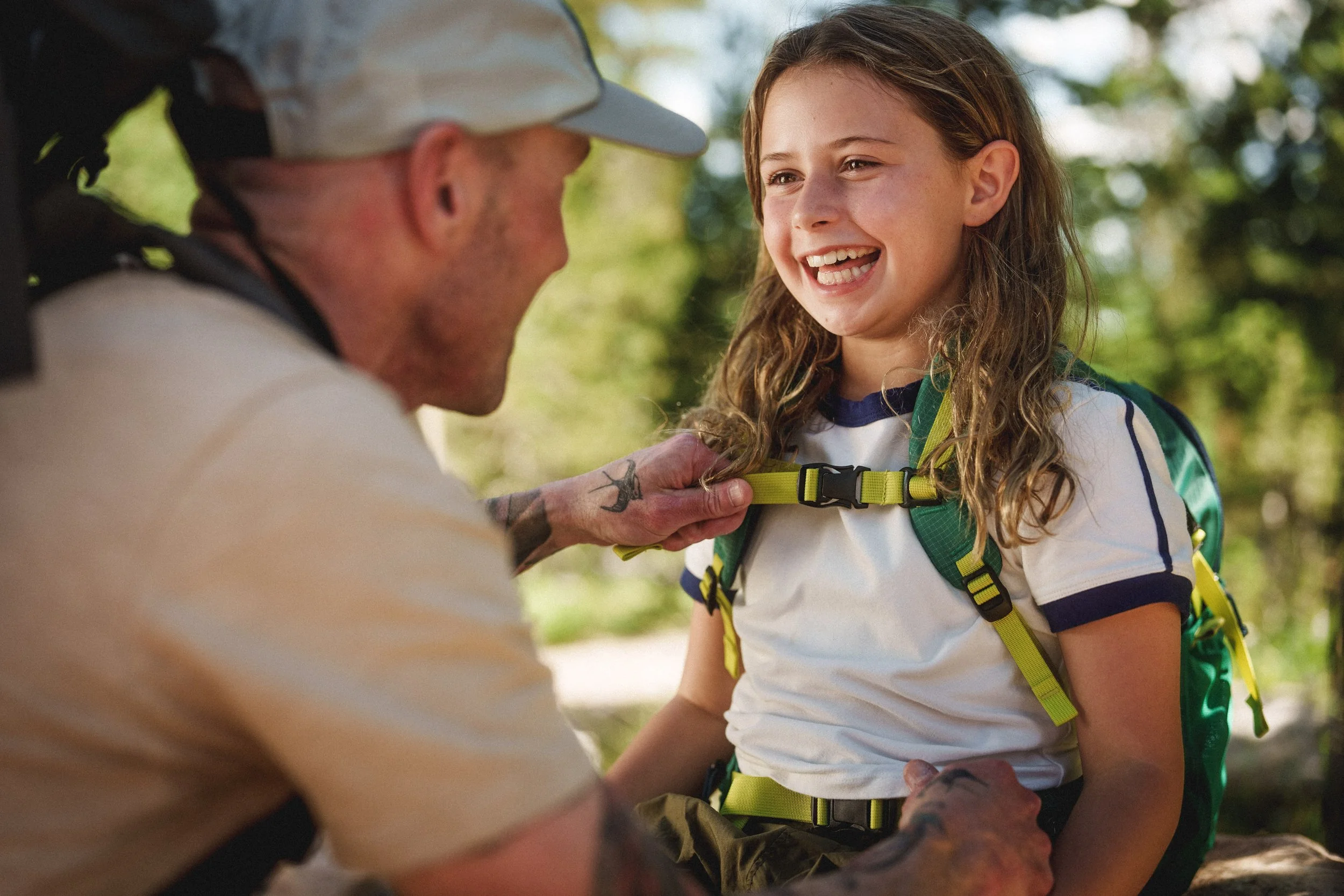 A young girl with curly hair wearing a white t-shirt with blue trim, smiling and laughing, wearing a yellow and green backpack, talking to a man with tattoos on his arms and neck, who is smiling and adjusting her backpack strap outdoors in a wooded area.