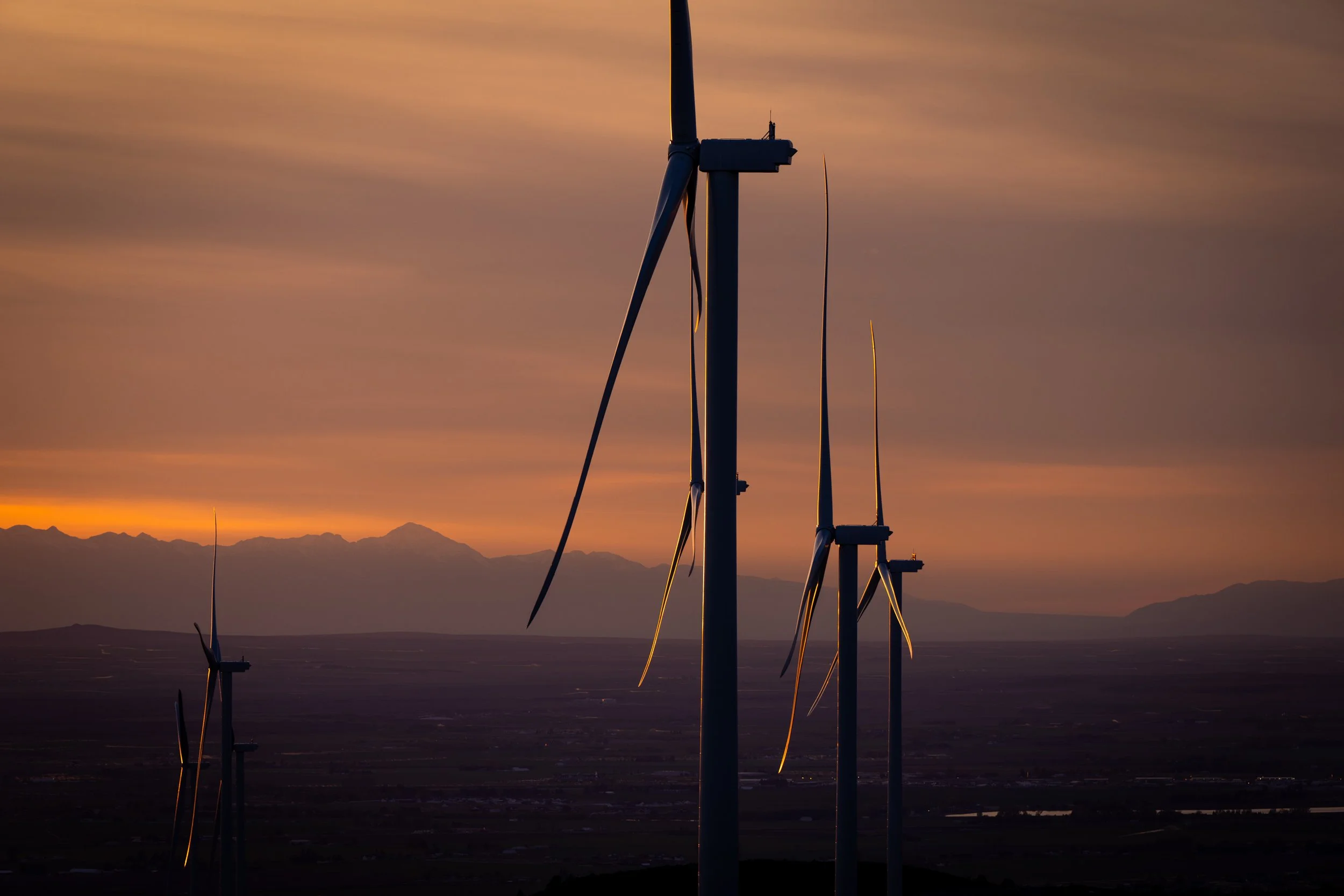 Silhouettes of wind turbines at sunset or sunrise with a mountain range in the background.