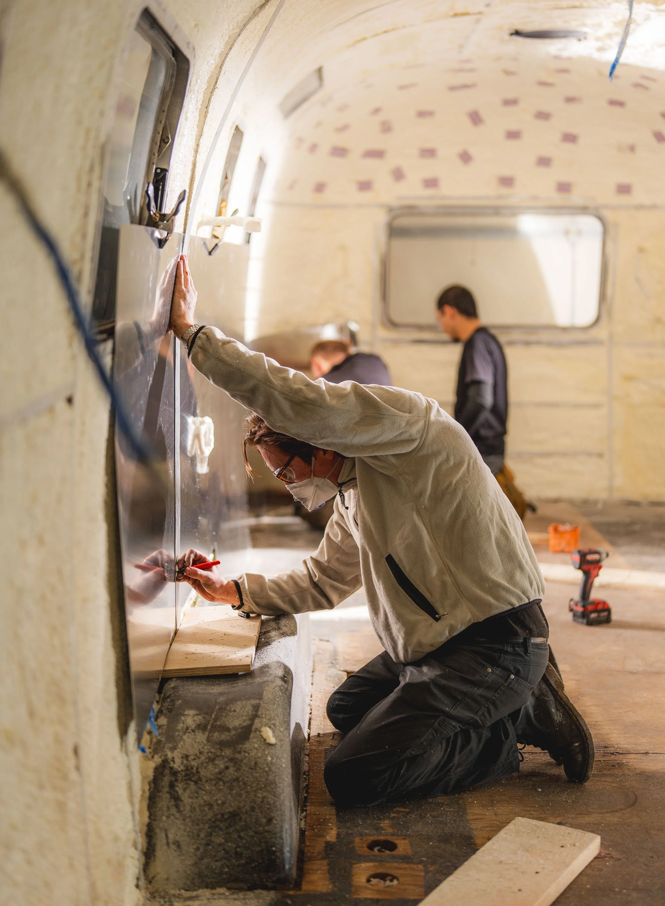 Person kneeling and marking a wall with a marker while working on renovation, with tools on the floor and other people in the background.