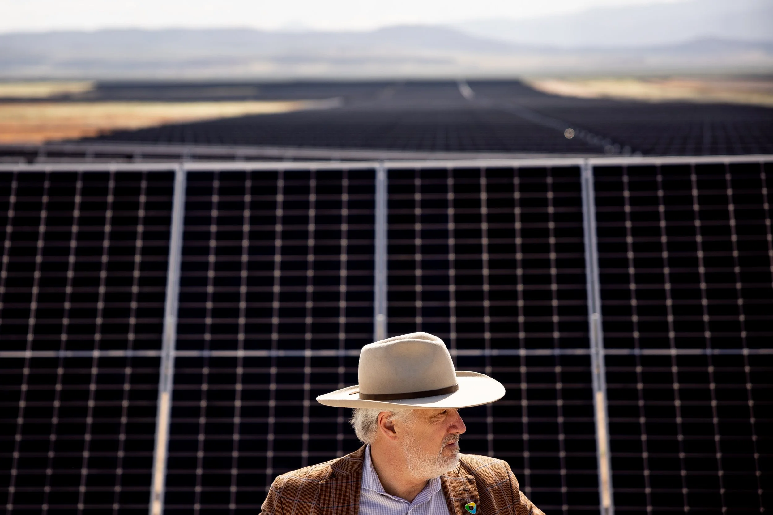 Senior man with a large hat and plaid jacket standing in front of solar panels in a sunny desert landscape.