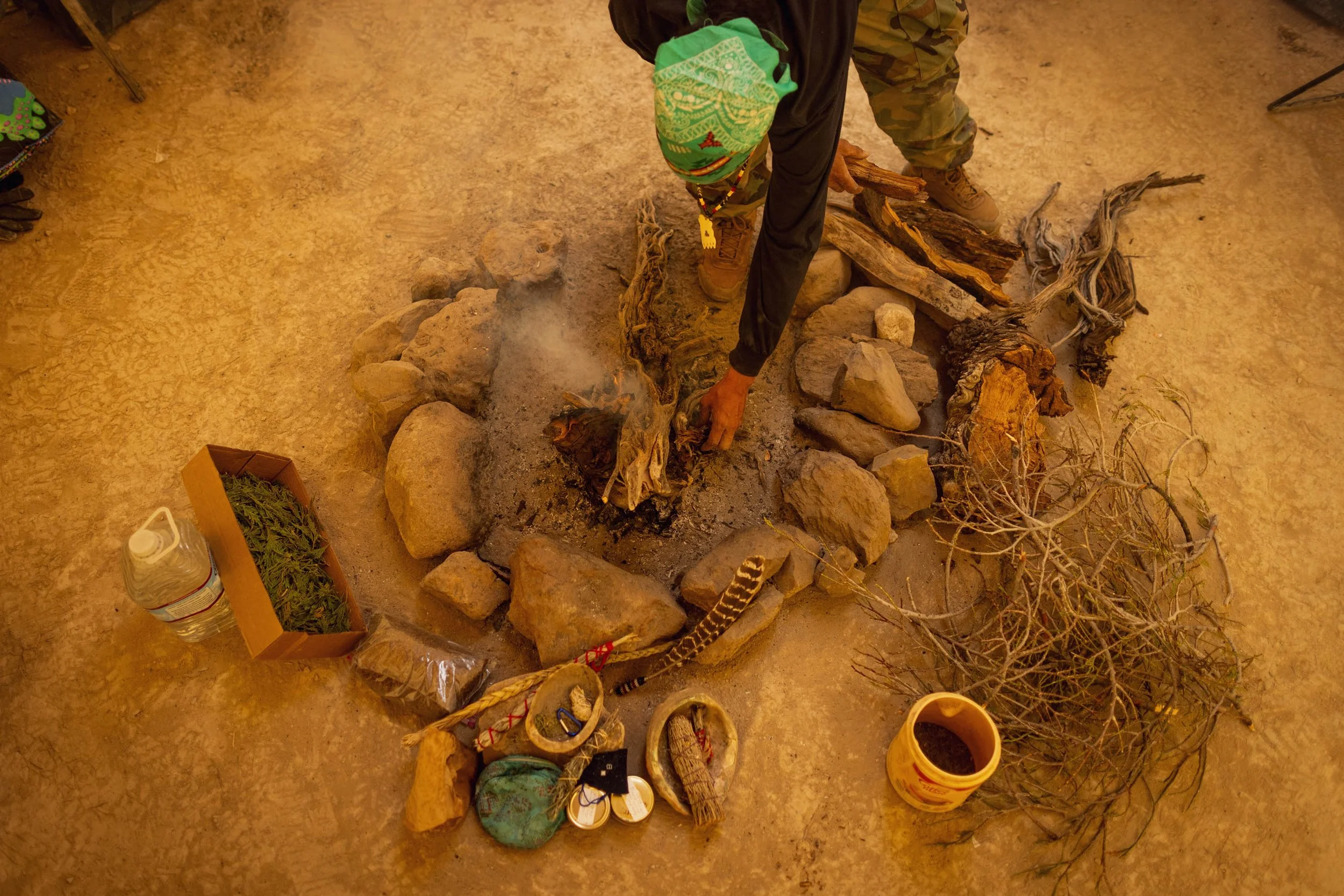 Person gathering herbs or plants around a small campfire made of logs and rocks, with various natural and man-made objects placed nearby.