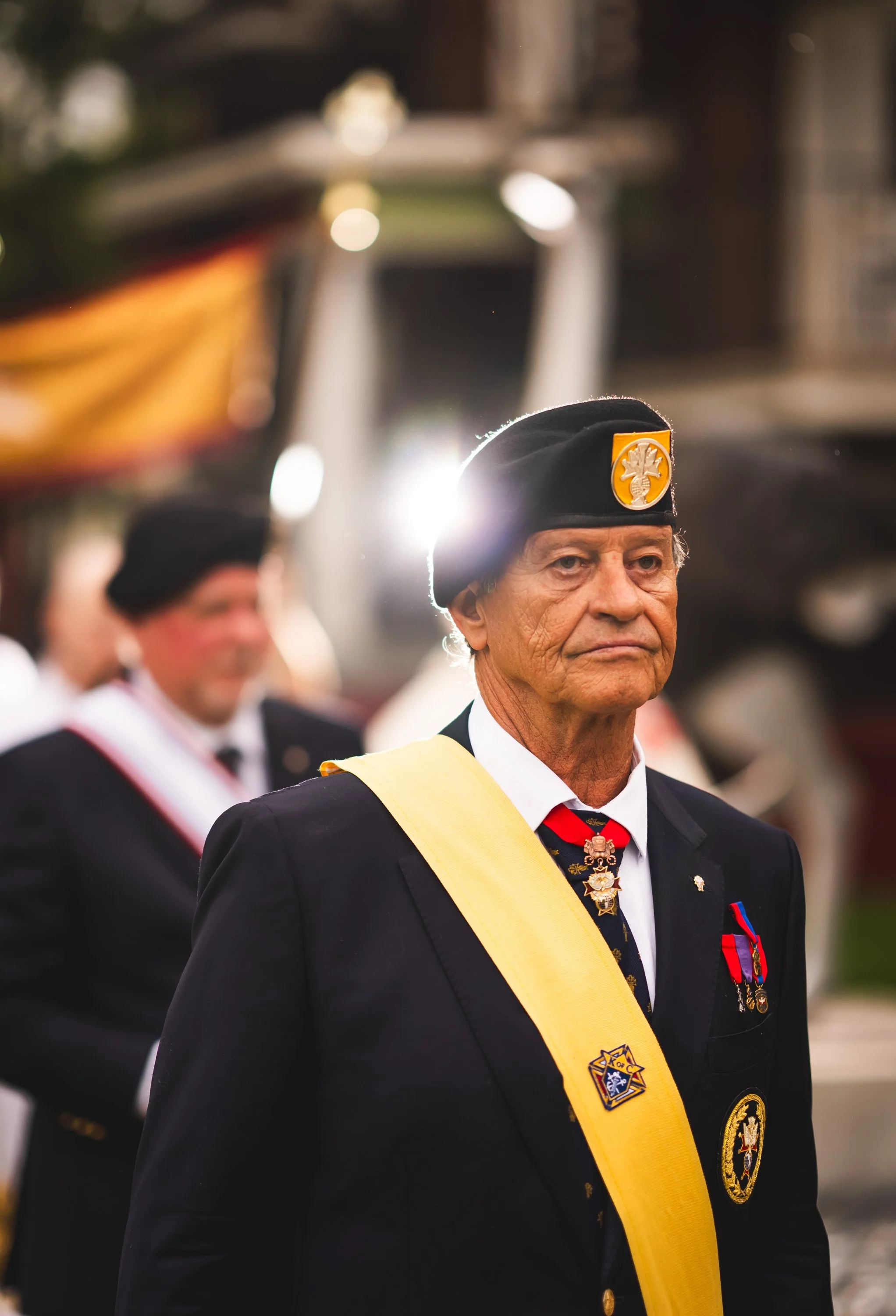 An elderly man dressed in formal military attire, wearing medals and a ribbon, standing in a parade with other veterans, some of whom are also dressed in similar uniforms and hats.