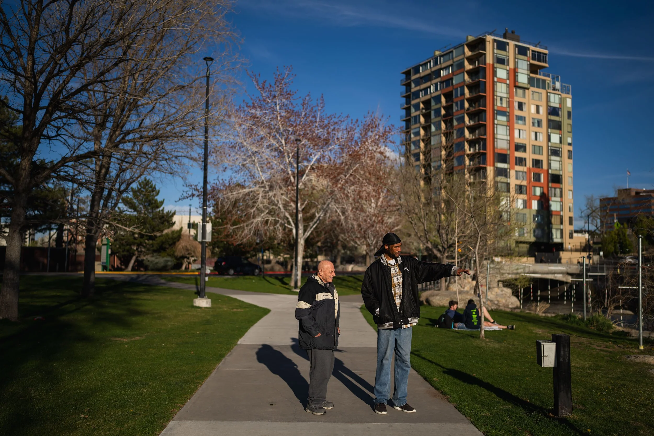 Two men stand on a sidewalk in a park, one pointing off in the distance, with leafless trees and a tall apartment building in the background, against a blue sky.