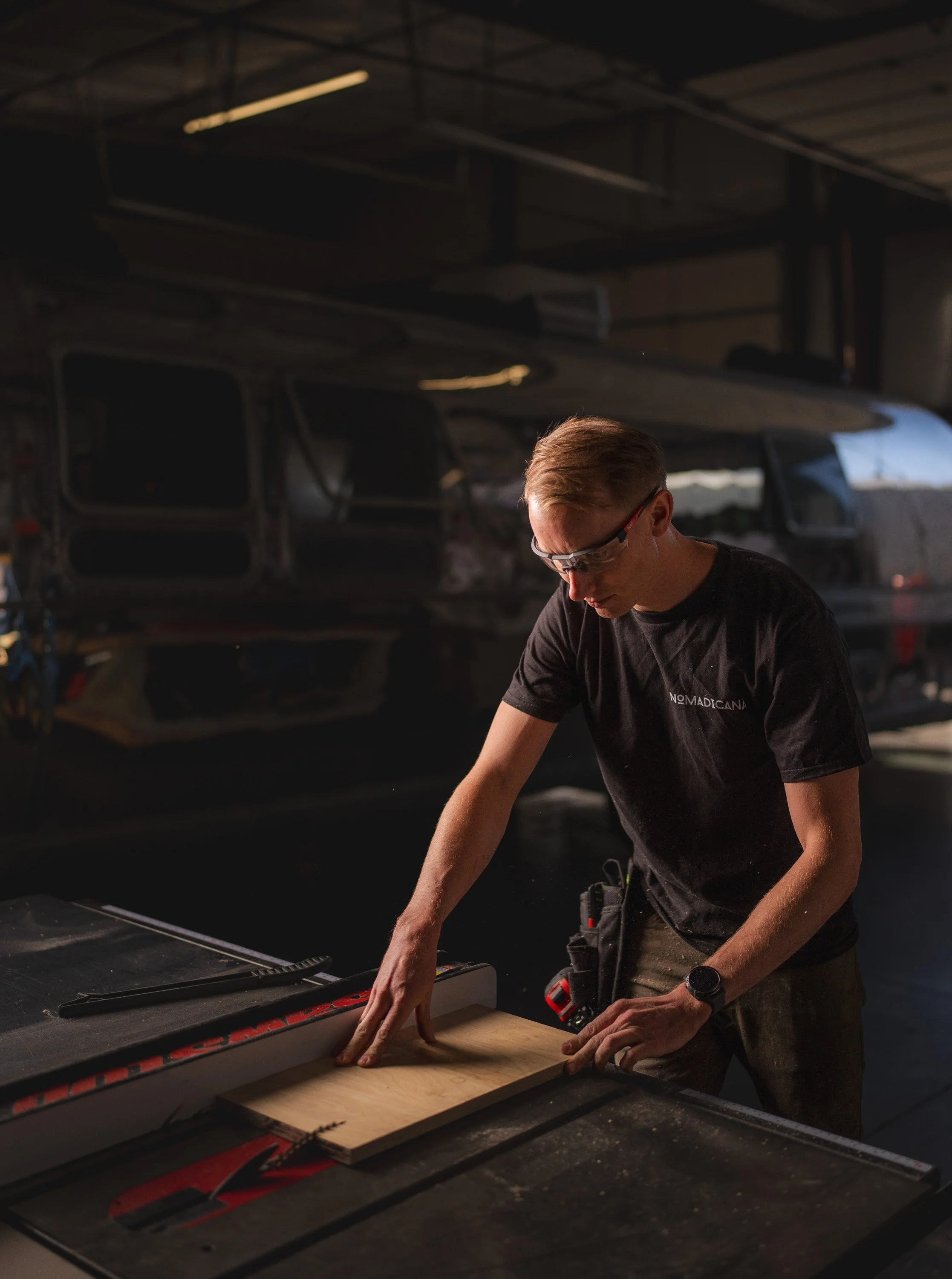 A person working in a workshop, measuring a piece of wood on a table saw. In the background, there is a partially assembled or disassembled airplane in a hangar.
