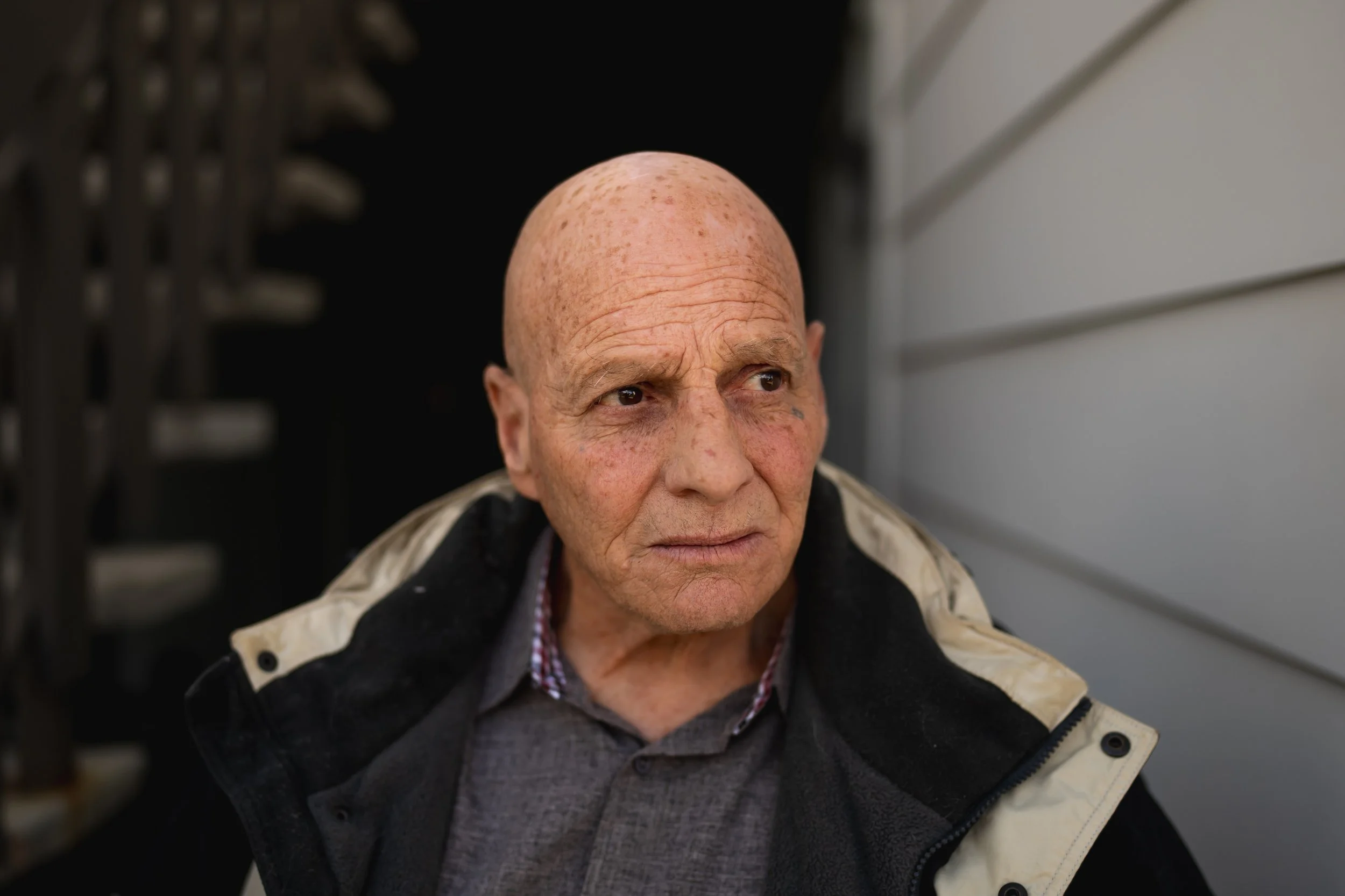 Close-up of an elderly bald man with a serious expression, wearing a grey shirt and a black and beige jacket, standing outdoors next to a house with white siding and a staircase in the background.